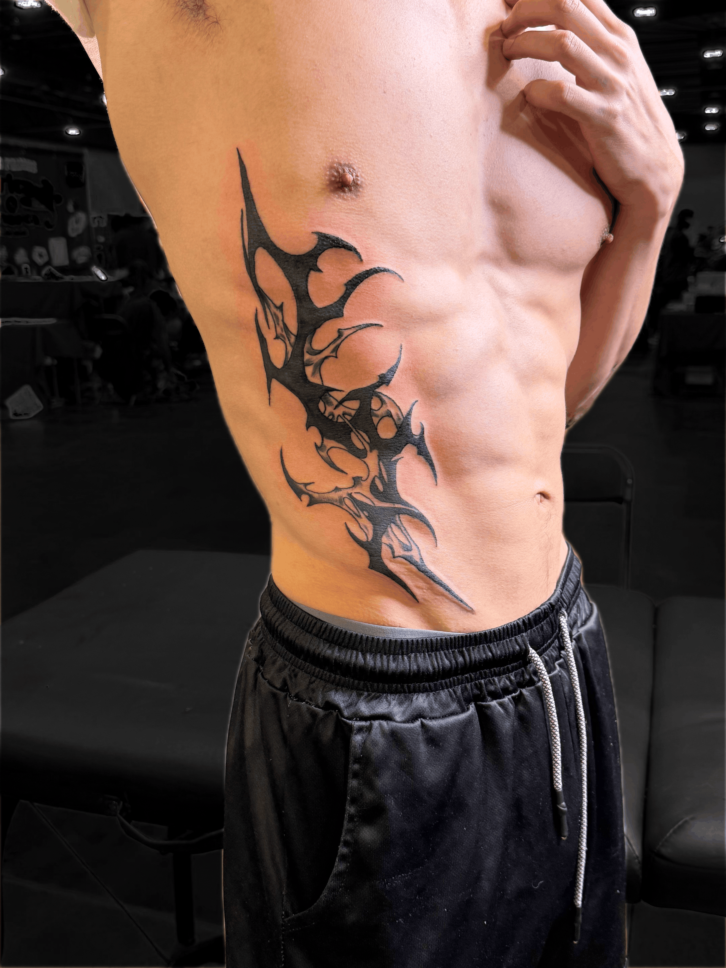 Close-up of a shirtless person with a large black cyber tattoo across their side and abdomen, wearing black shorts. Work by Hannah.