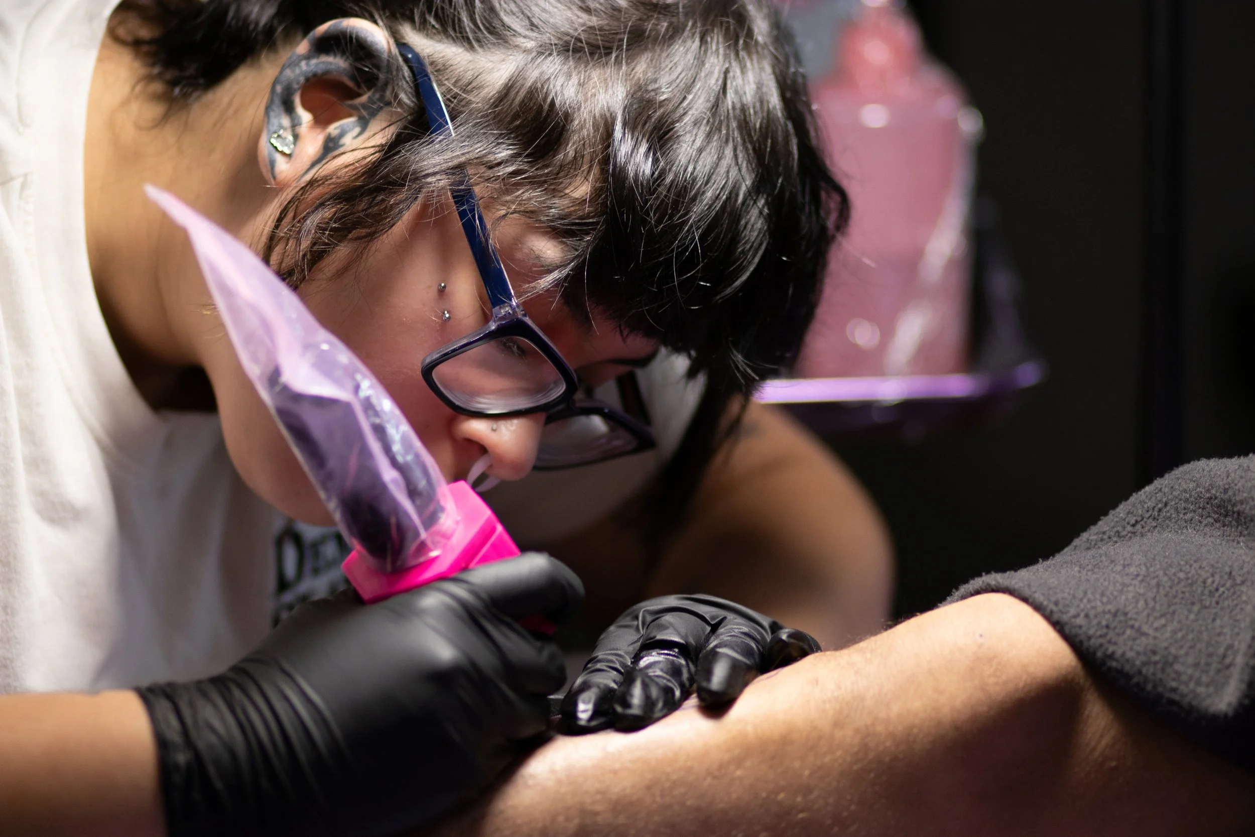 Tattoo artist Sury wearing glasses and black gloves tattooing a client’s arm with a purple bagged tattoo machine.