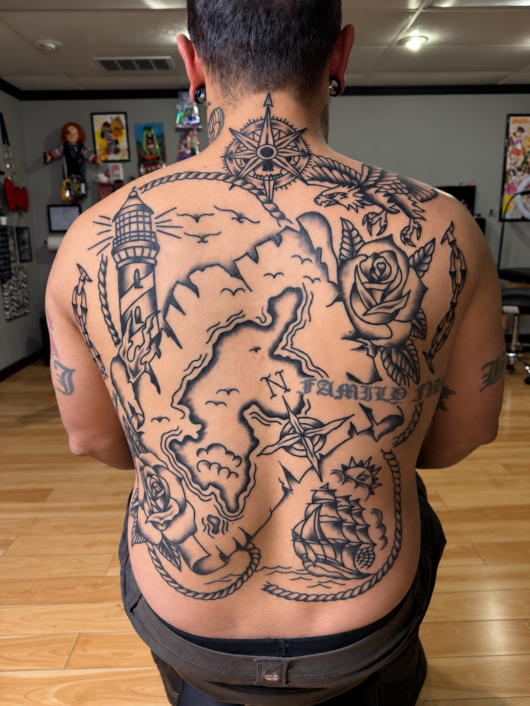 Person displaying an extensive nautical tattoo covering their back, featuring a compass, map, lighthouse, eagle, rose, anchor, and sailing ship. By Ruben