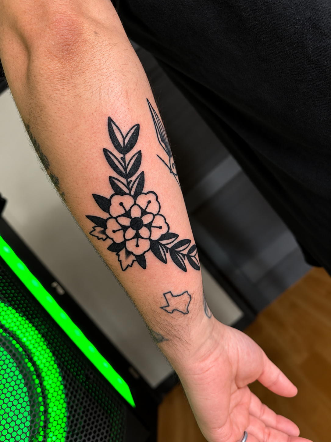 Close-up of a forearm featuring a black floral tattoo with leaves and a small Texas outline tattoo near the wrist, work done by Ruben.
