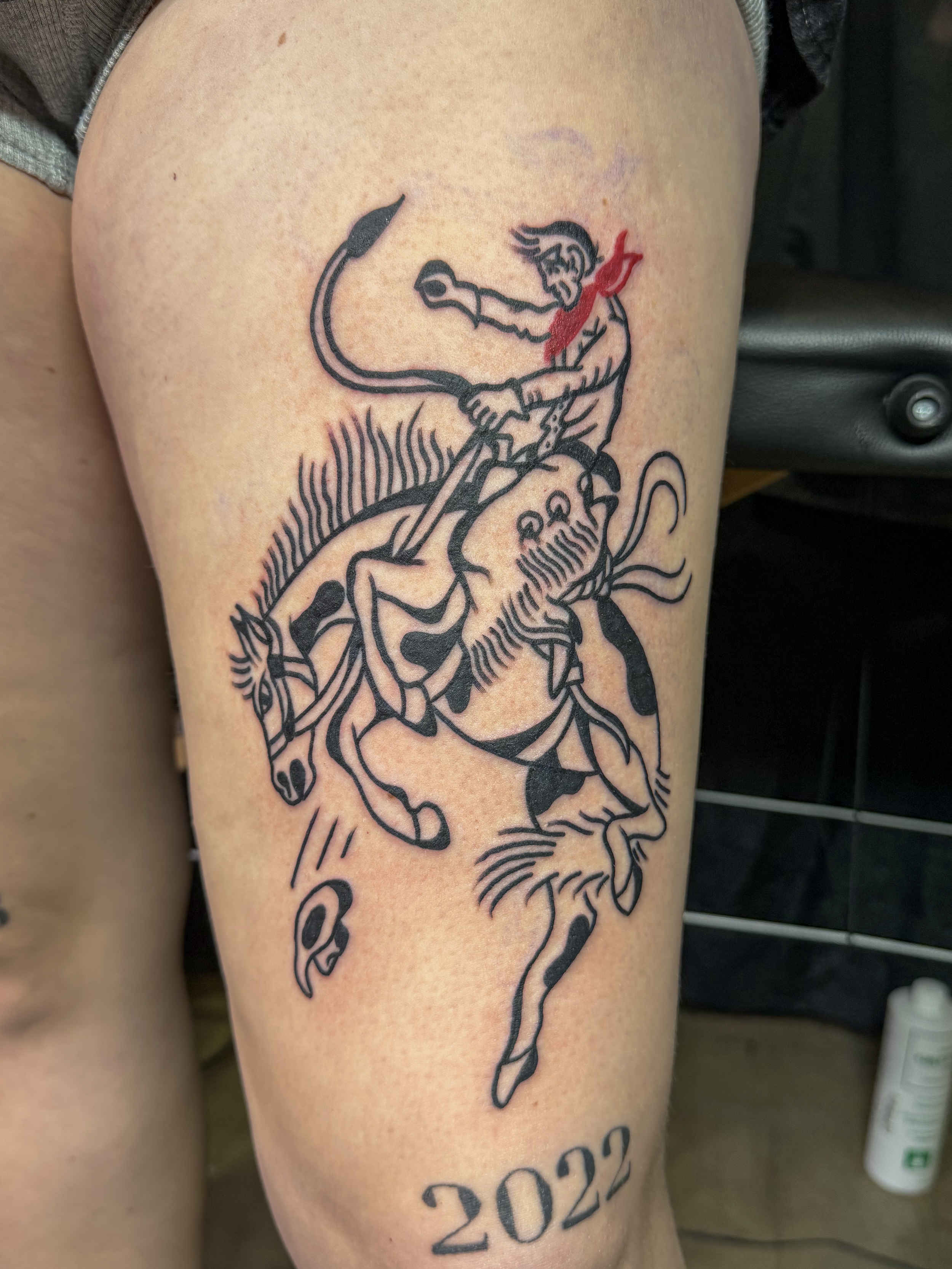 Close-up of a black-and-red cartoon-style tattoo featuring a seated cowboy bucking a bronco horse. By Ruben.
