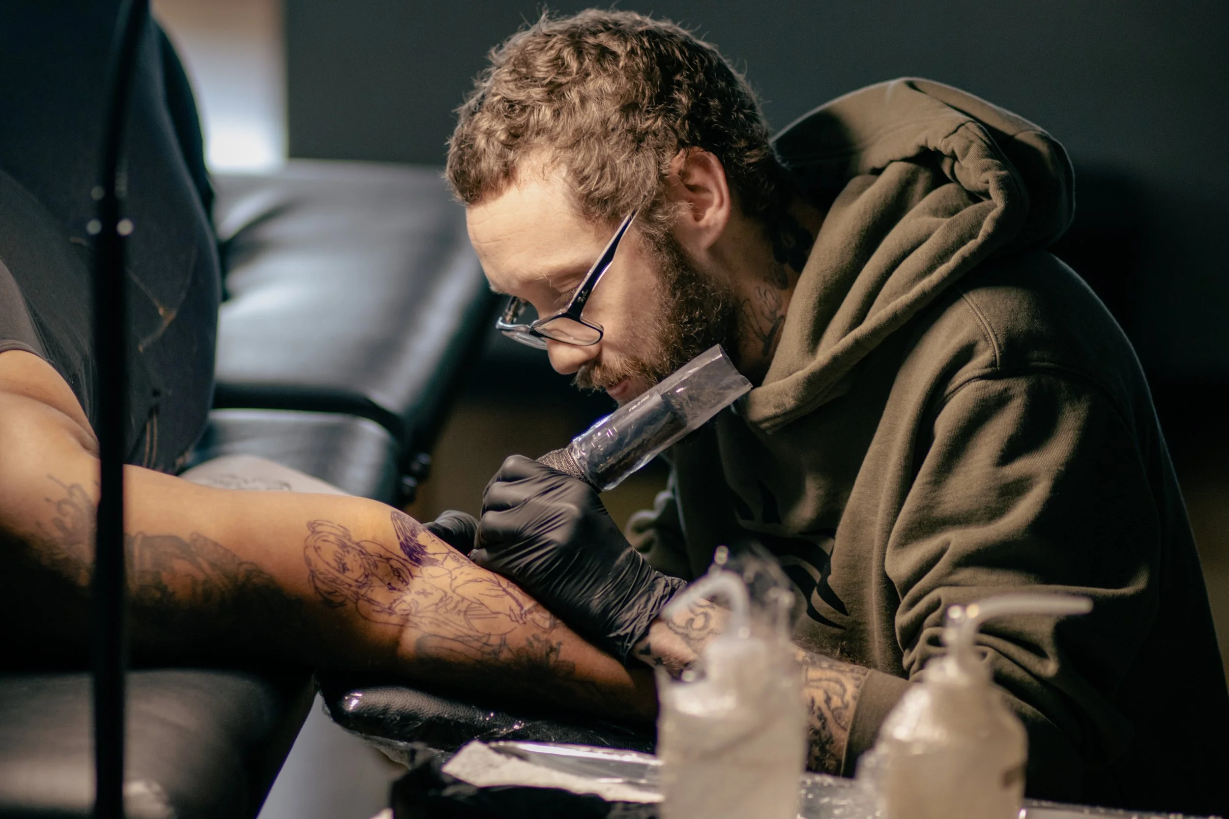 Tattoo artist Pancho wearing glasses and gloves tattooing a client's arm in a studio.
