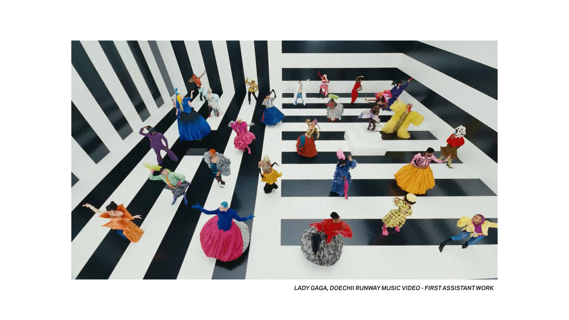 Large group of dancers in colorful costumes performing on a black-and-white striped geometric set