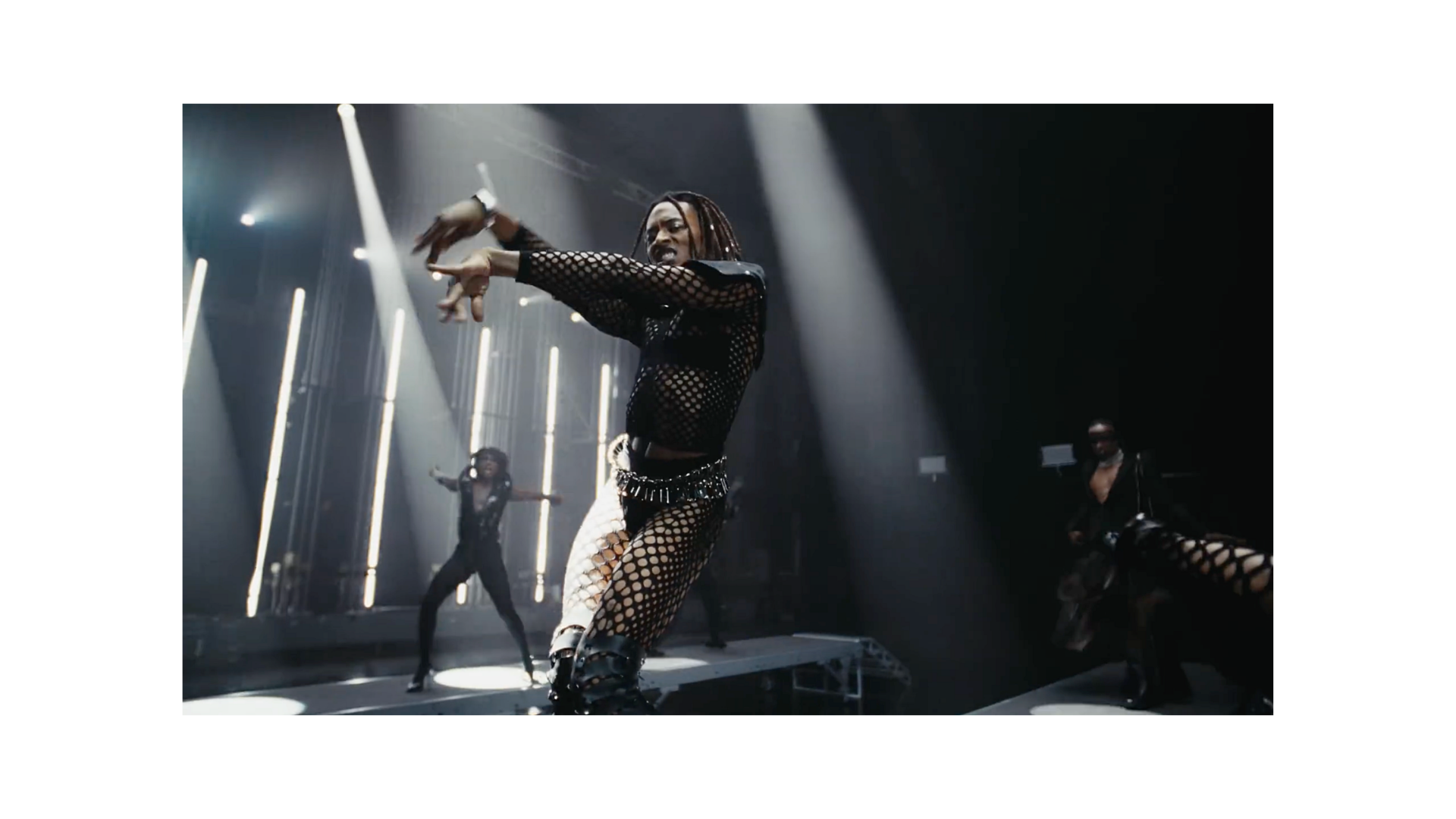 Singer in a black fishnet outfit performing choreography on a brightly lit concert stage with backup dancers