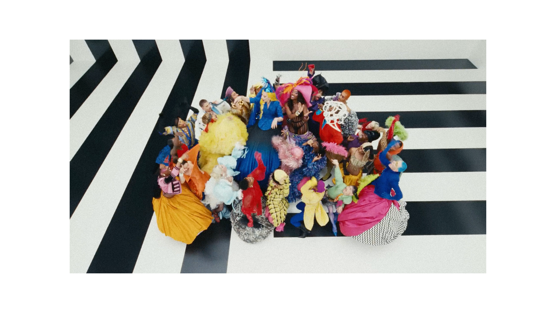Large group of people in colorful costumes and elaborate hats posing on a black-and-white geometric set