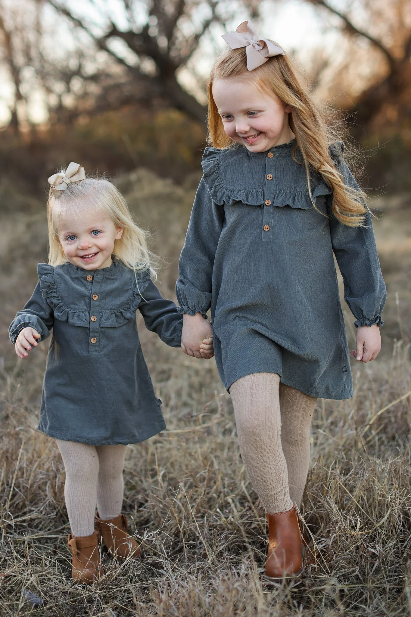 Two smiling young sisters in matching gray dresses and bows holding hands outdoors in a grassy field