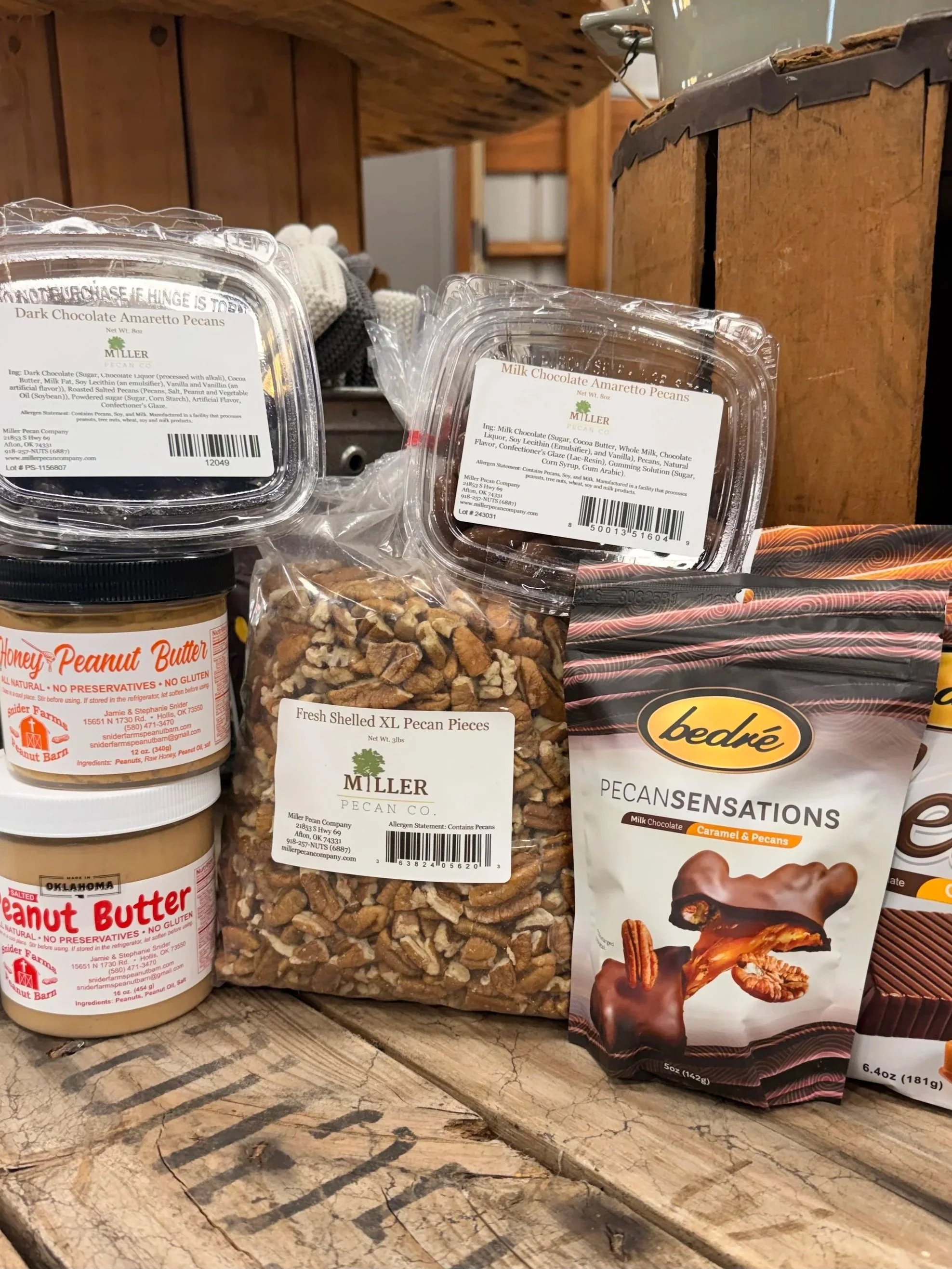 Assorted pecan products, including chocolate-covered pecans, shelled pecan pieces, peanut butter, and pecan candy packages on a rustic wooden table.