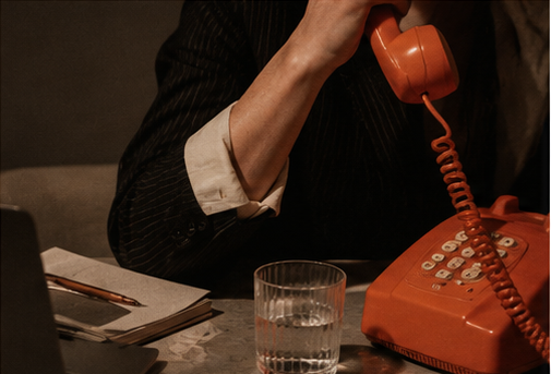 Person holding a red corded telephone handset beside a glass of water and notebook