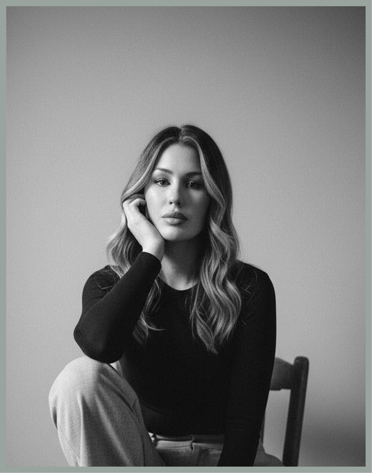 Black-and-white portrait of a woman with long wavy hair sitting in a chair with her hand resting against her face