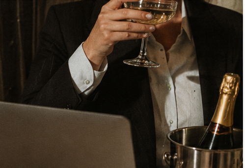 Businessman in a suit holding a champagne glass beside a laptop and bottle of champagne in an ice bucket