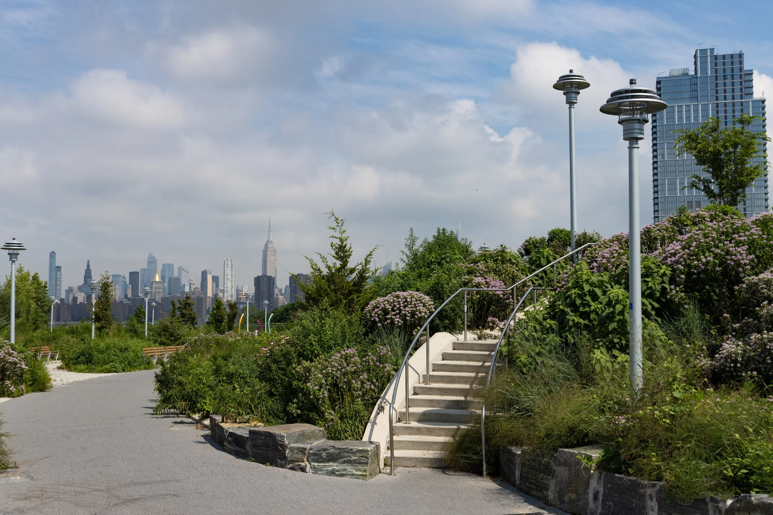 Landscaped urban park with stairs, modern lampposts, and a Manhattan skyline featuring the Empire State Building