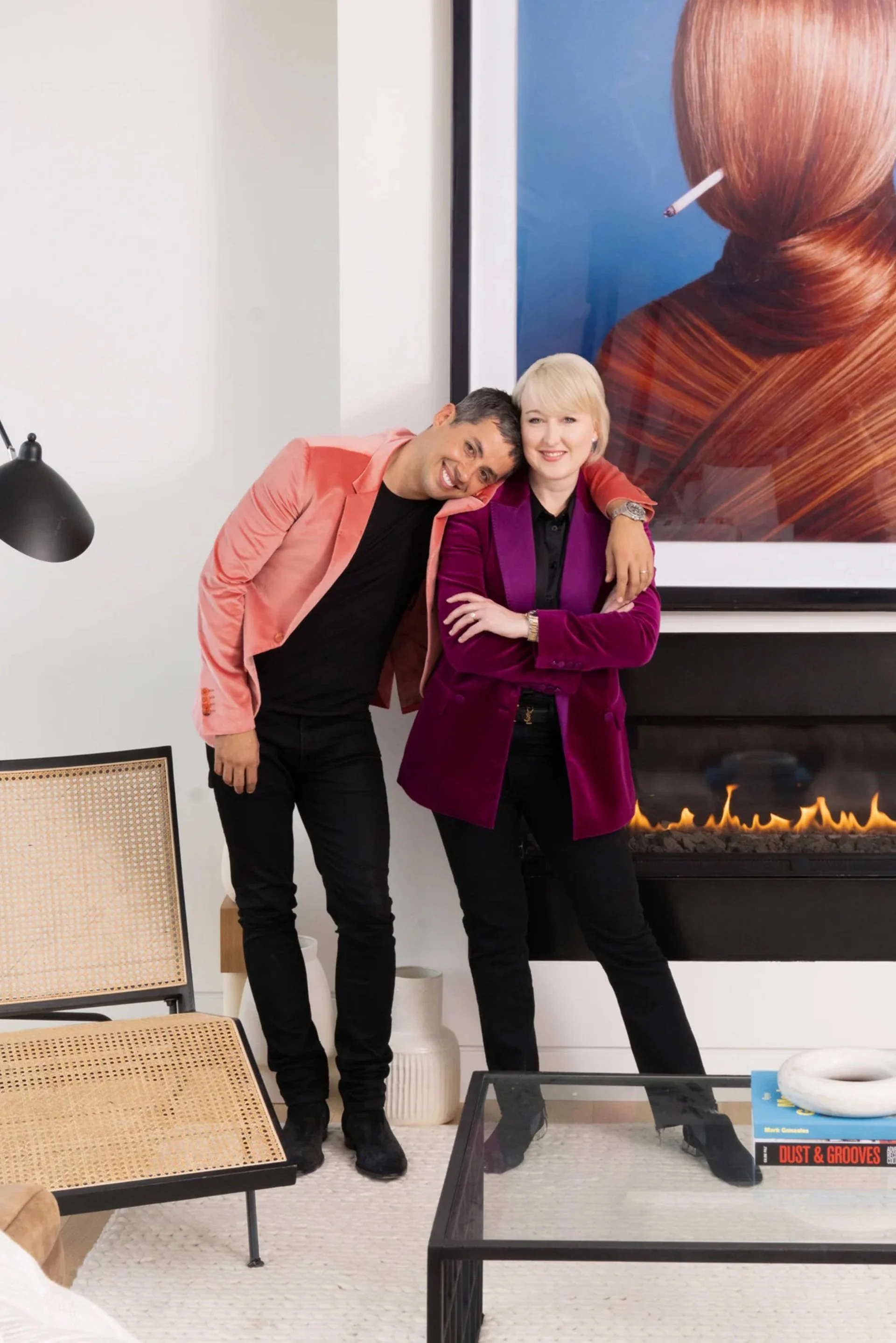 Smiling man in a pink blazer and woman in a purple blazer posing together in a modern living room with a fireplace and colorful artwork