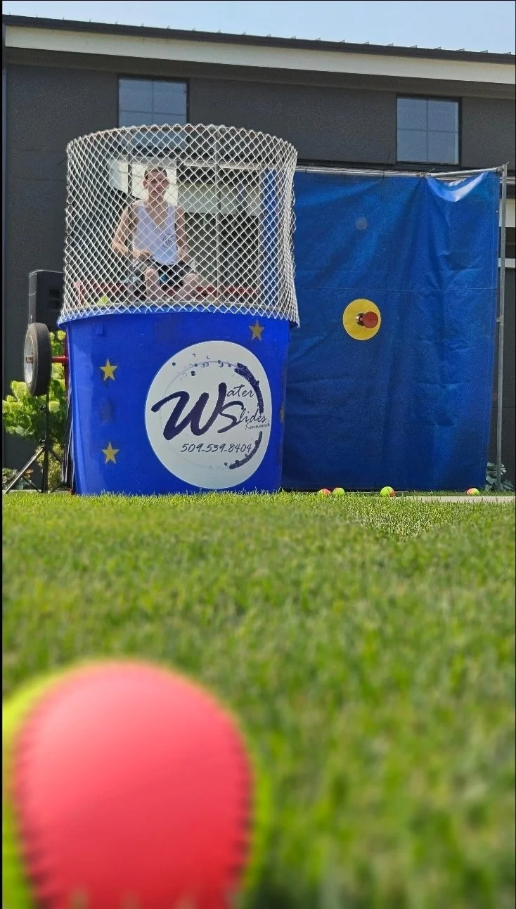 Person seated inside the blue Dunk Dynasty dunk tank behind the safety net, with the throwing target visible at an outdoor event.