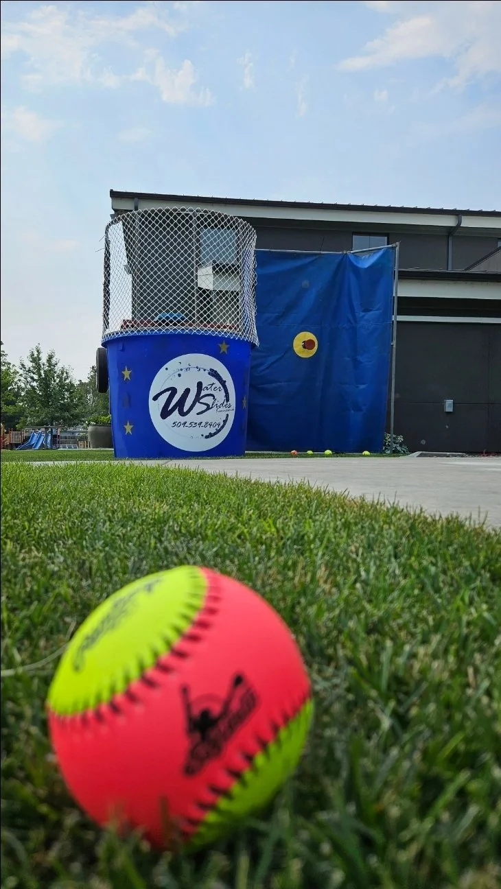 Bright neon softball on the grass in front of the Dunk Dynasty blue dunk tank and target, available for Tri-Cities party rentals.