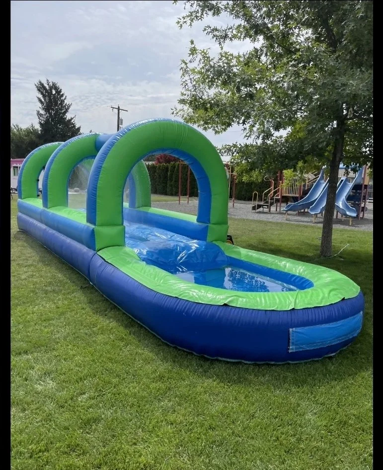 Slip Happens green and blue inflatable slip-and-slide with splash pool set up on a grassy lawn near playground equipment.