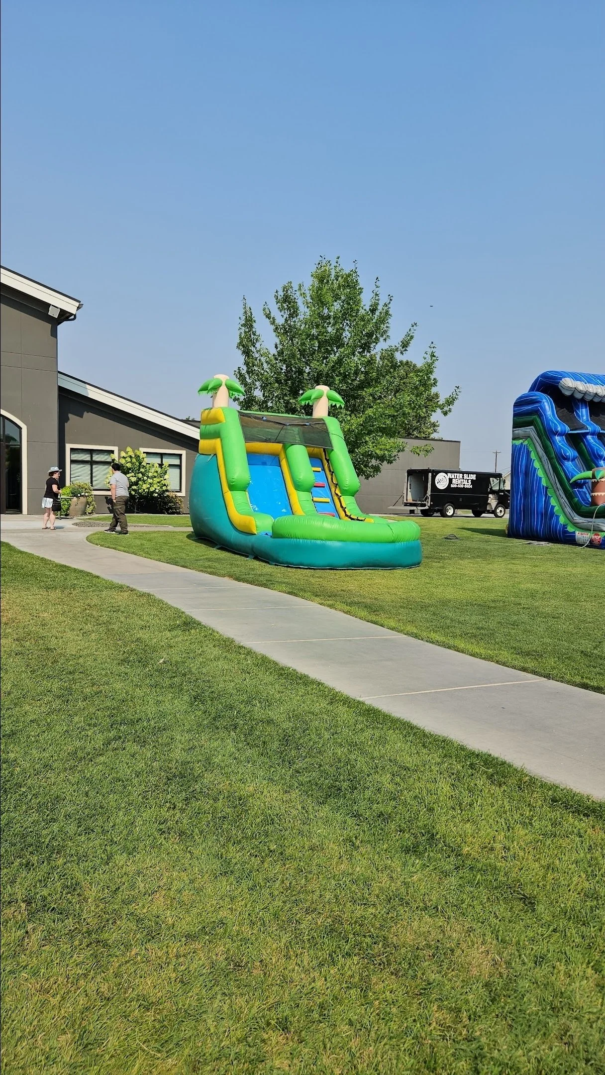 The Tiki inflatable waterslide photographed from across a grassy lawn, showing the outdoor setup and surrounding yard space.
