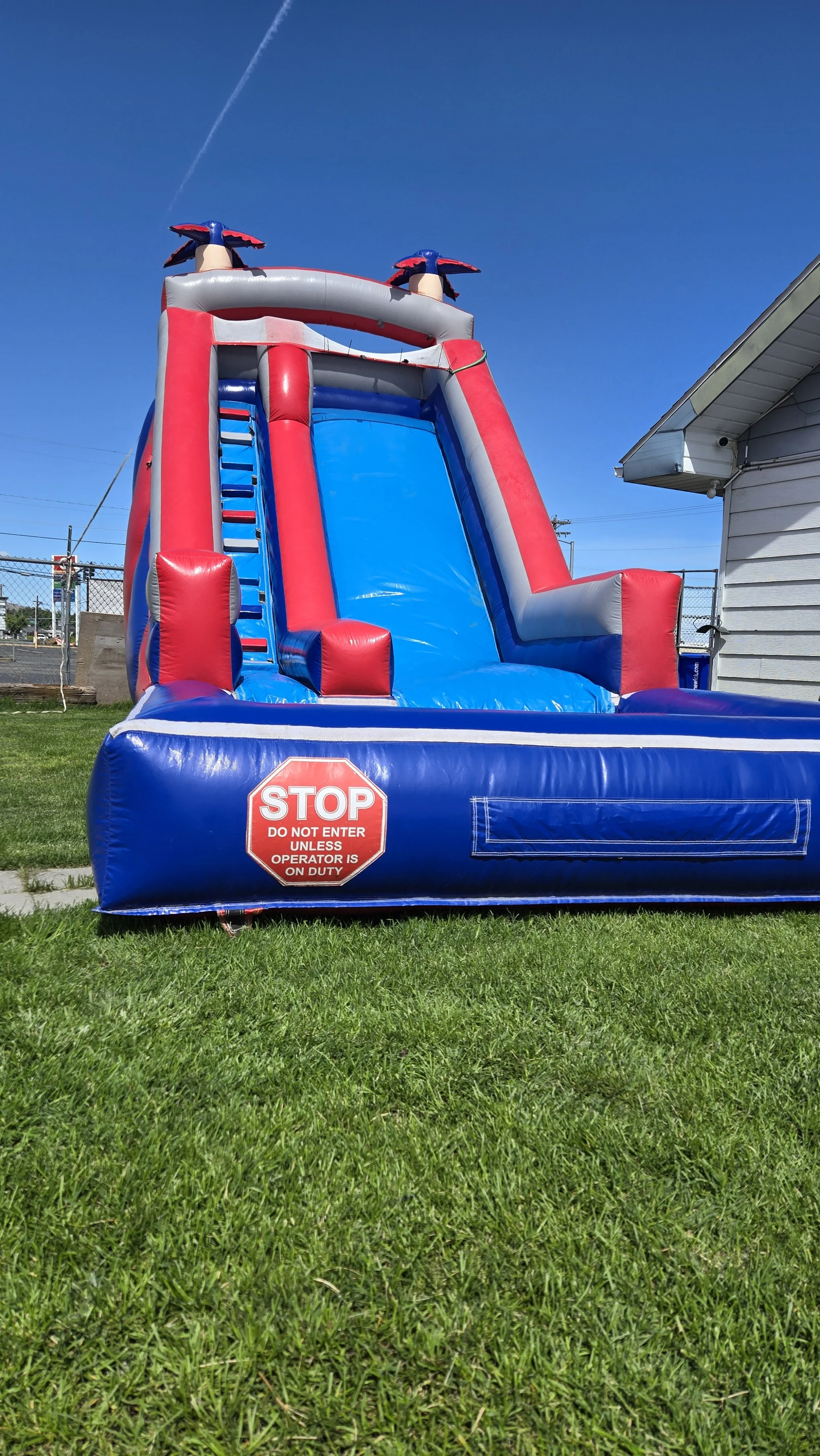 Red White N' Cool inflatable waterslide rental standing on a sunny grassy lawn near a commercial building, available for parties in Richland, Washington.