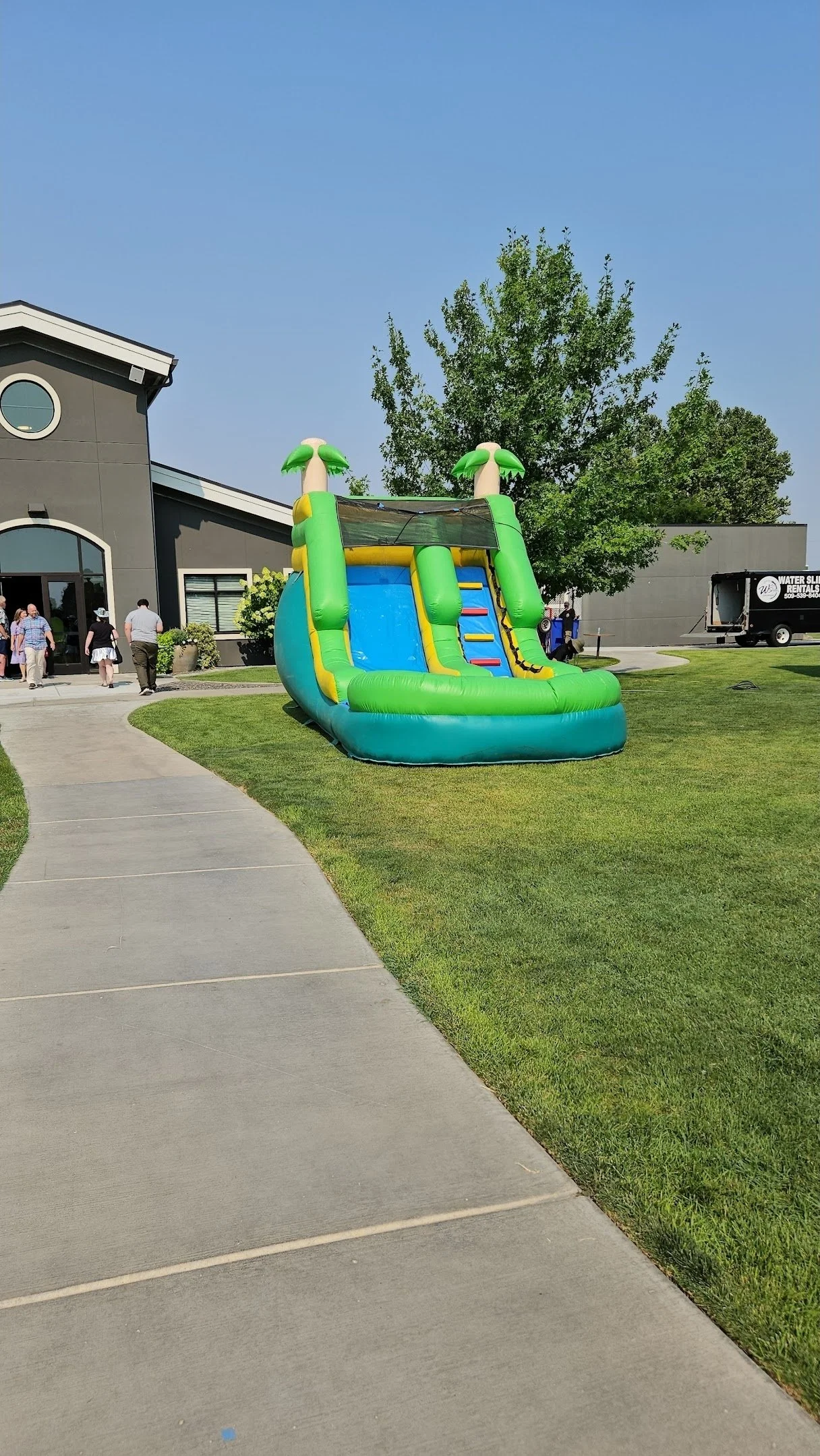 Angled view of The Tiki green tropical waterslide showing its inflatable structure and palm-tree decorations beside a grassy yard.