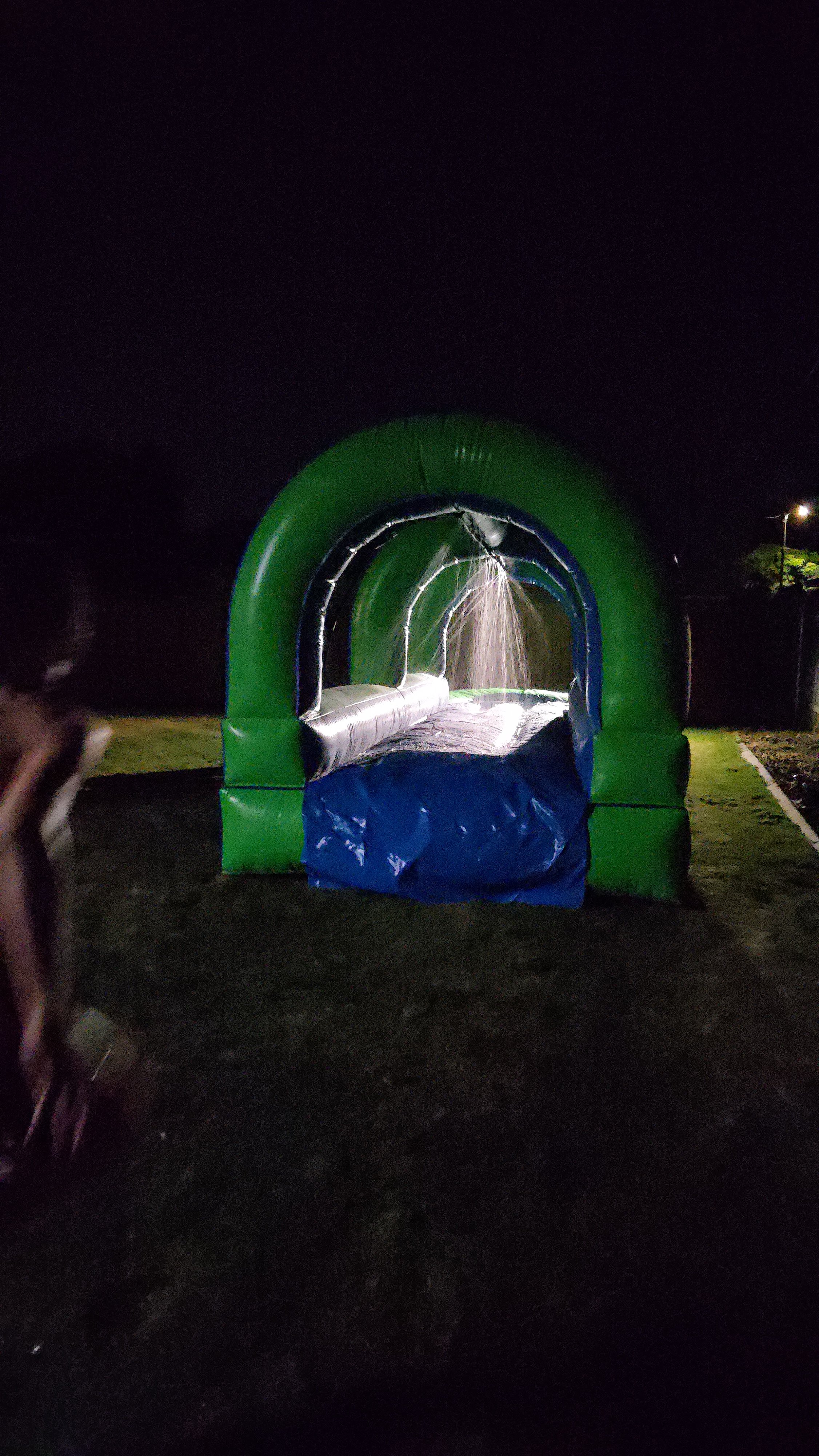 Slip Happens 28-foot inflatable slip-and-slide illuminated at night, showing the water jets spraying along the green and blue slide.