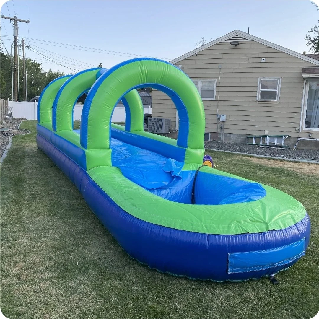 Angled view of the Slip Happens 28-foot inflatable slip-and-slide with green entrance arch and blue splash pool on a backyard lawn.