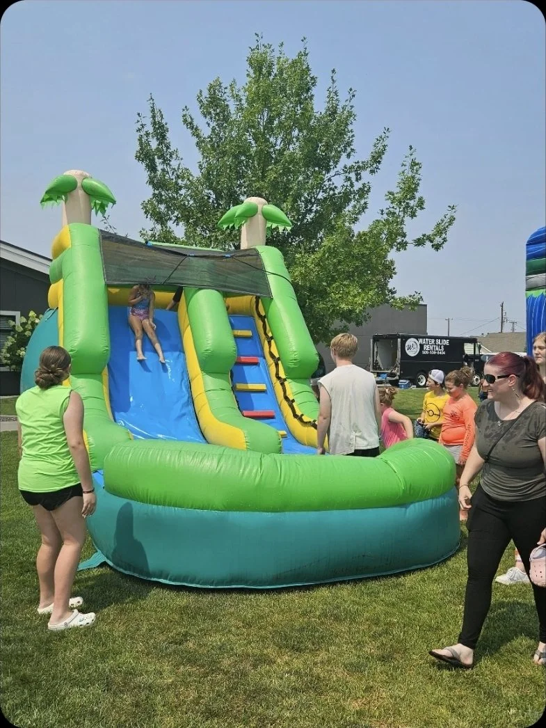 Children enjoying The Tiki green tropical-themed inflatable waterslide with palm-tree decorations at an outdoor summer event.