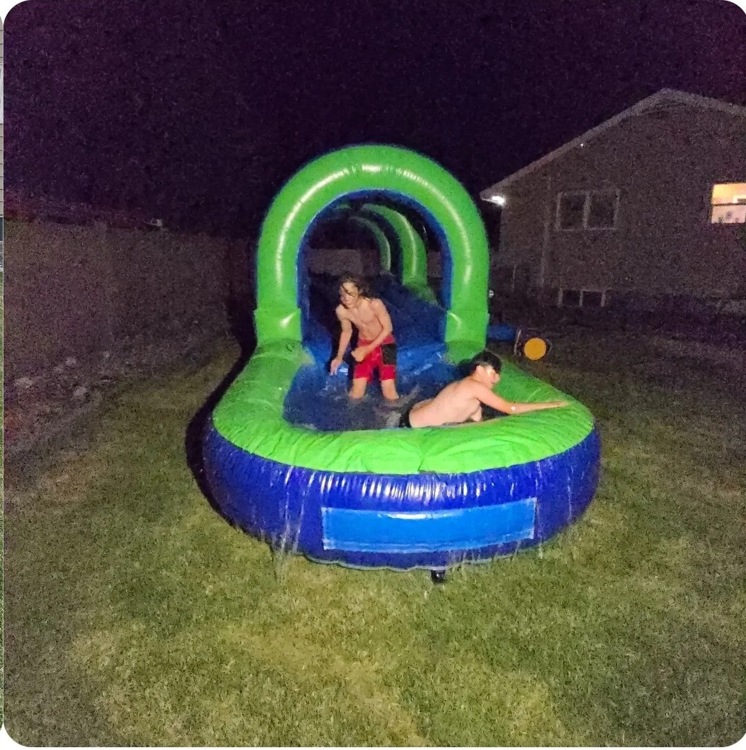 Two boys enjoying the Slip Happens green and blue inflatable slip-and-slide and splash pool outdoors at night.