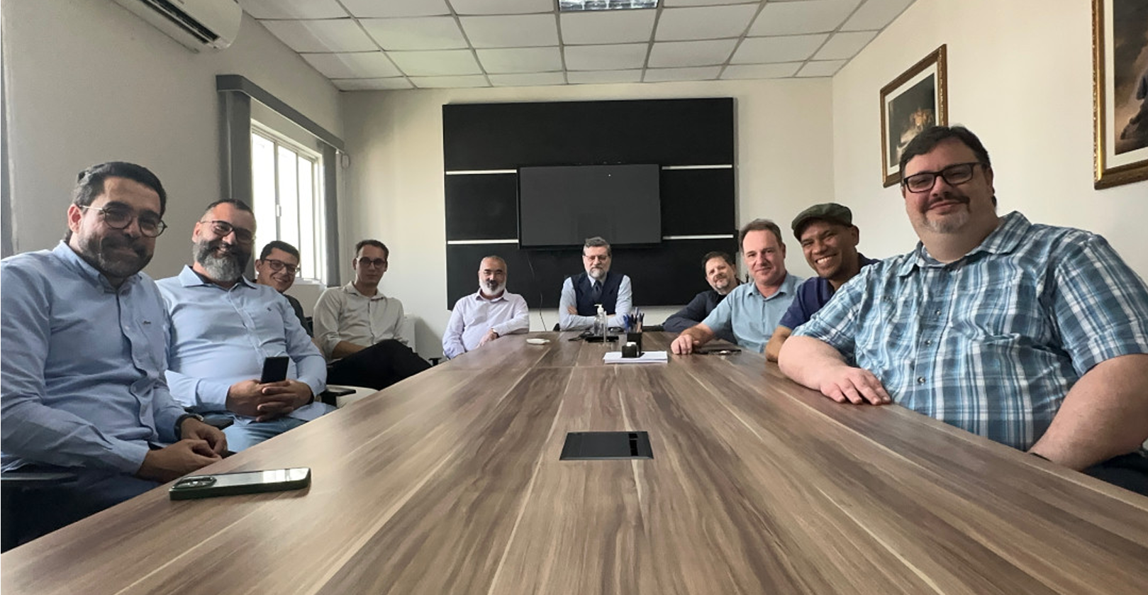 Group of twelve men sitting around a conference table in a meeting room, smiling at the camera.