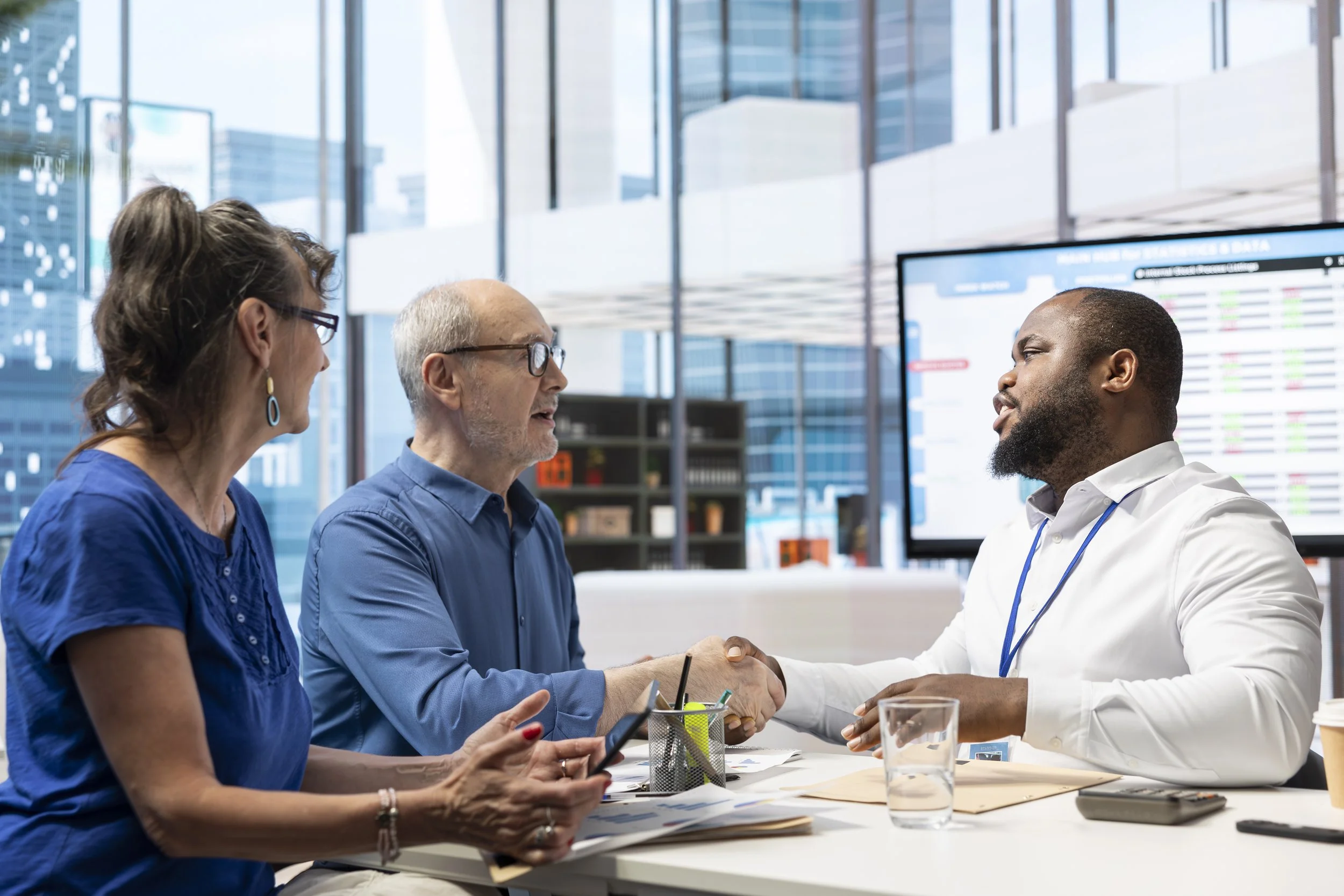 Three business professionals in an office shaking hands. Two are women, and one is a man. They are sitting at a table with documents, a glass of water, and a smartphone. Large screens with data are visible in the background.