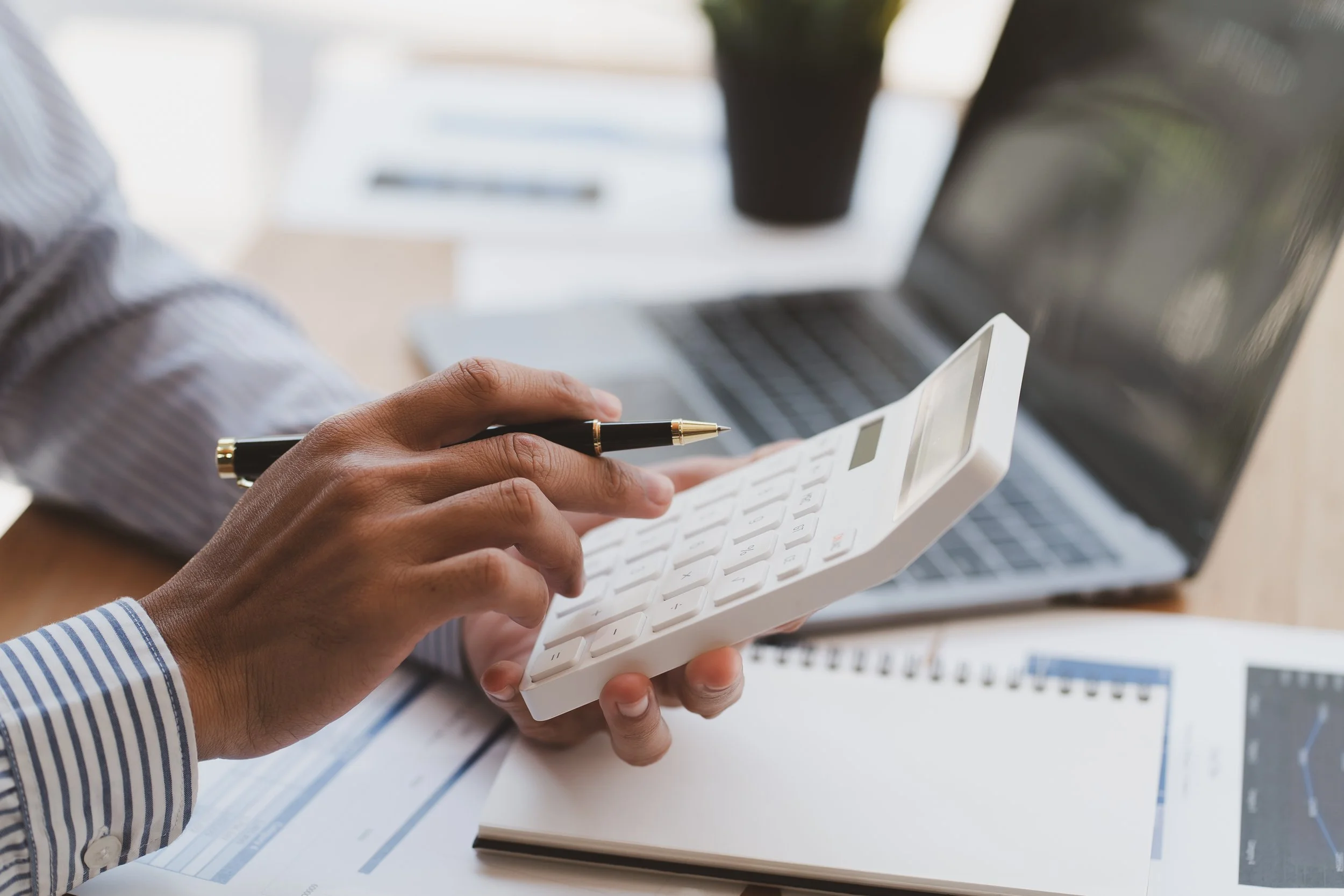 Person holding a calculator and a pen, with a laptop and documents on a desk in an office setting.
