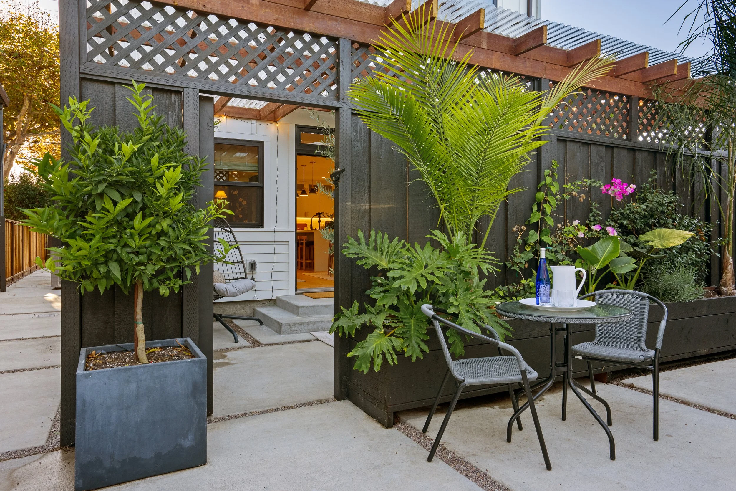 A modern backyard patio with lush green plants and trees, a round table with a blue water bottle and glass, two gray chairs, a black privacy fence, and a house with open door showing warm interior lighting.