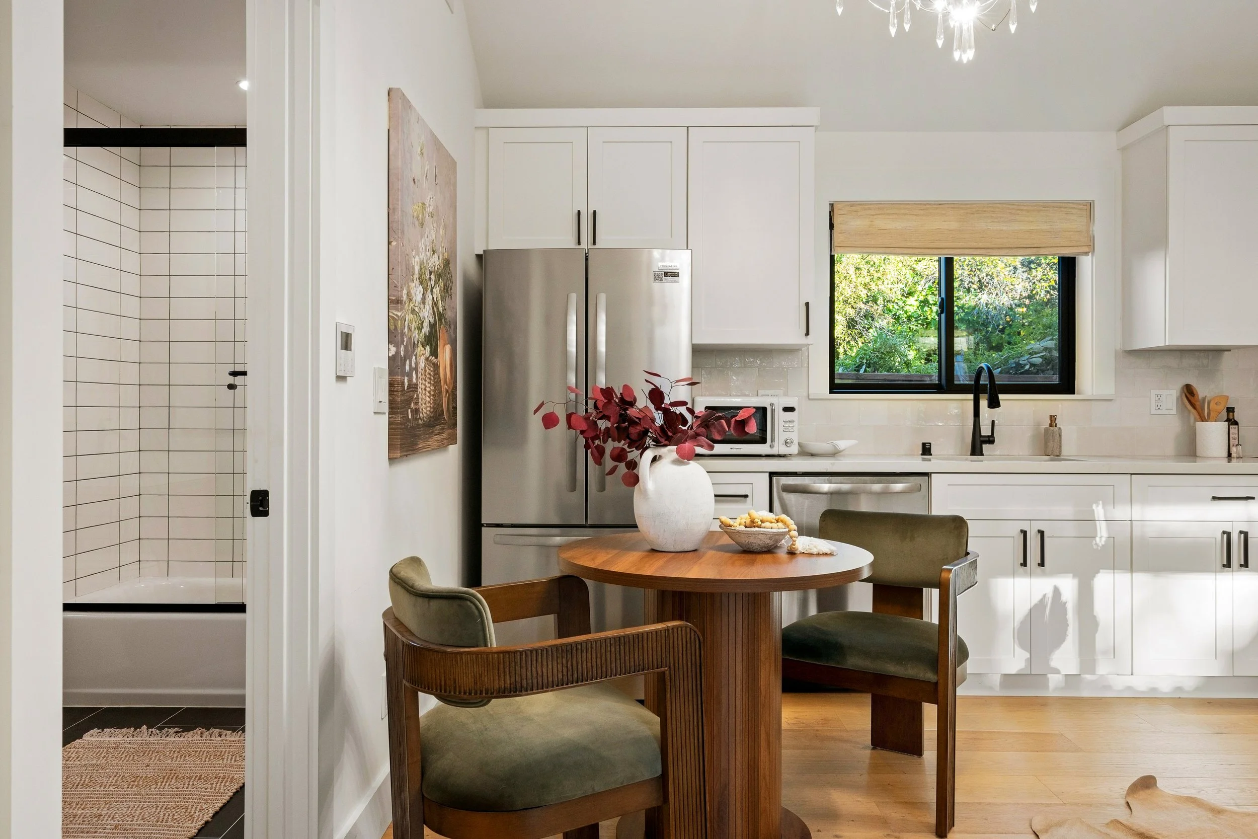 A modern kitchen with white cabinets, a stainless steel refrigerator, and a window with a beige shade. There is a round wooden dining table with chairs, a white vase with red leaves, and a small bowl of snacks. To the left, a doorway leads to a tiled
