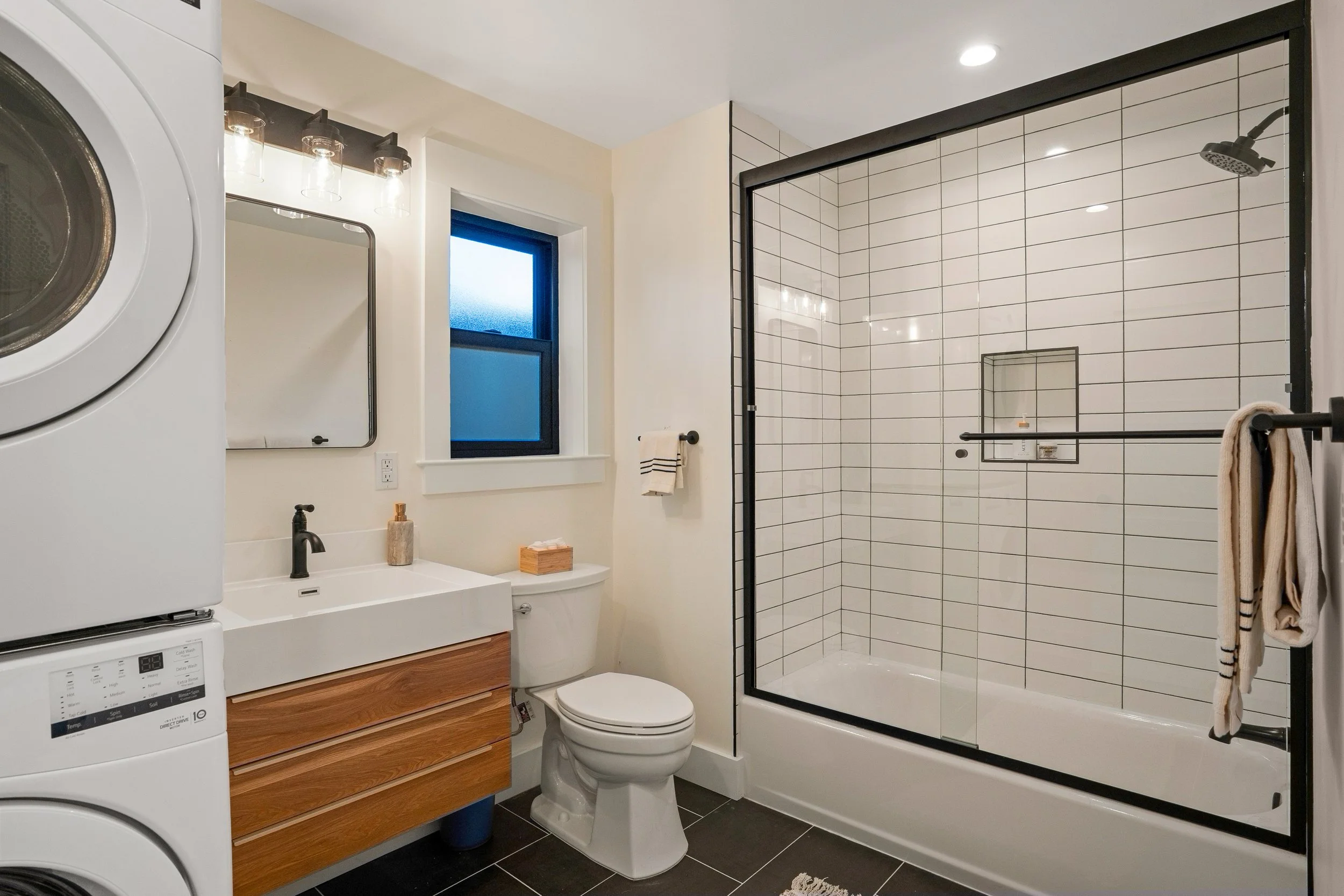Modern bathroom with a front-loading washer and dryer, a white vanity with a wooden drawer, a rectangular mirror, a frosted window, a toilet, a glass shower with black framing and white subway tiles, and a beige towel hanging on a black towel bar.