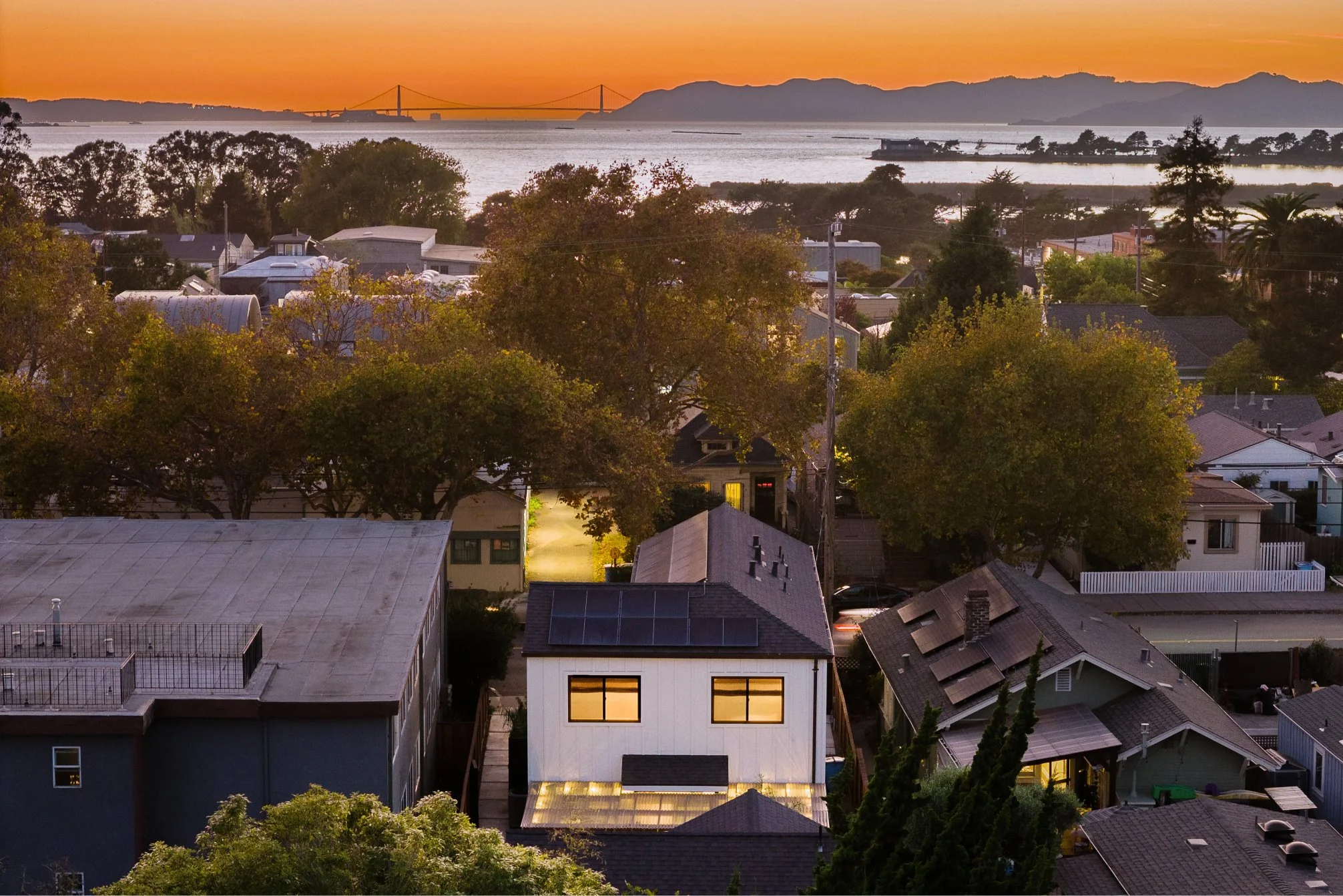 View of a residential neighborhood at sunset with houses, trees, and a backlit sky over the water and distant hills.