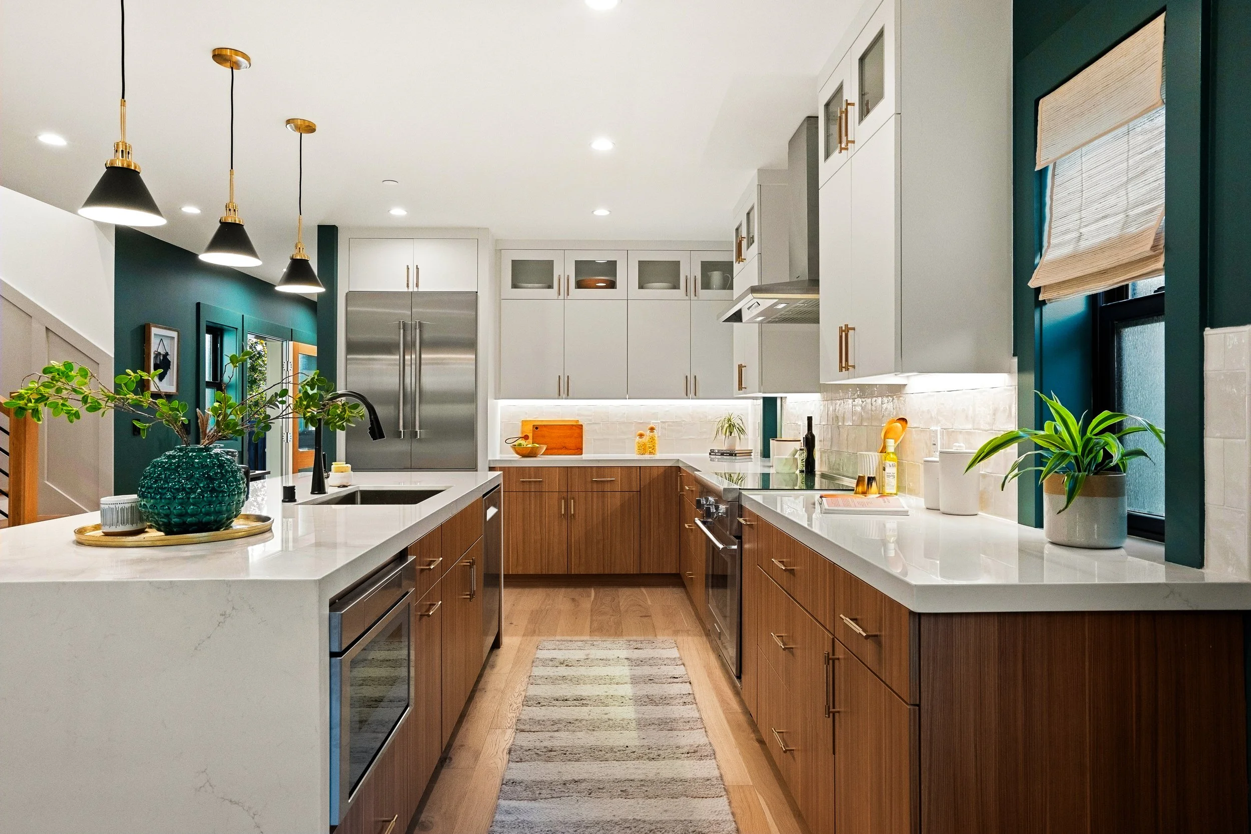 Modern kitchen with white and wood cabinets, stainless steel refrigerator, black pendant lights, green accent wall, and decorative plants on the white countertop.
