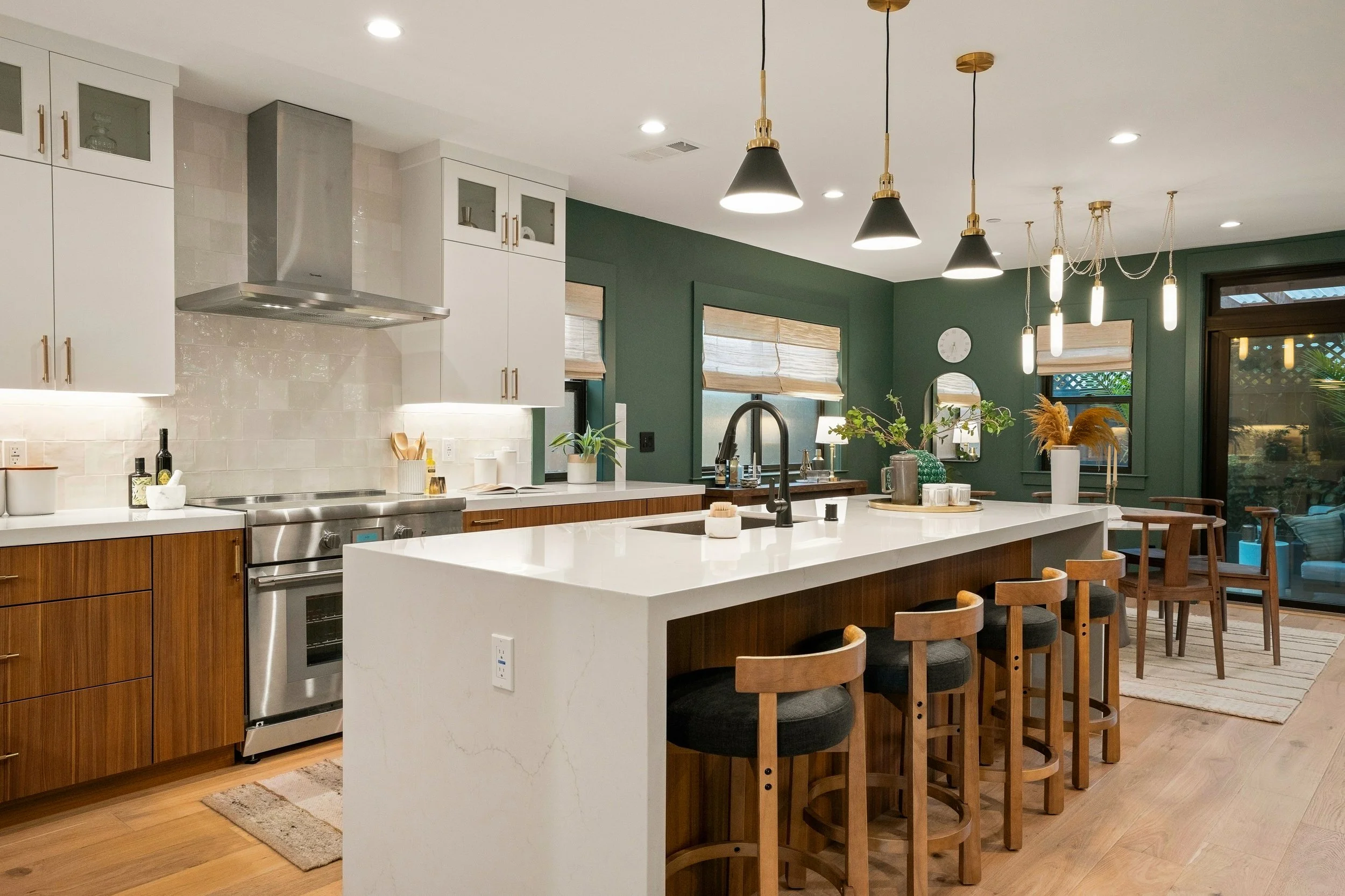 Modern kitchen with white and wood cabinetry, stainless steel appliances, and a large white island with seating. Green accent wall with windows, hanging pendant lights, and a dining area with wooden chairs and decor.