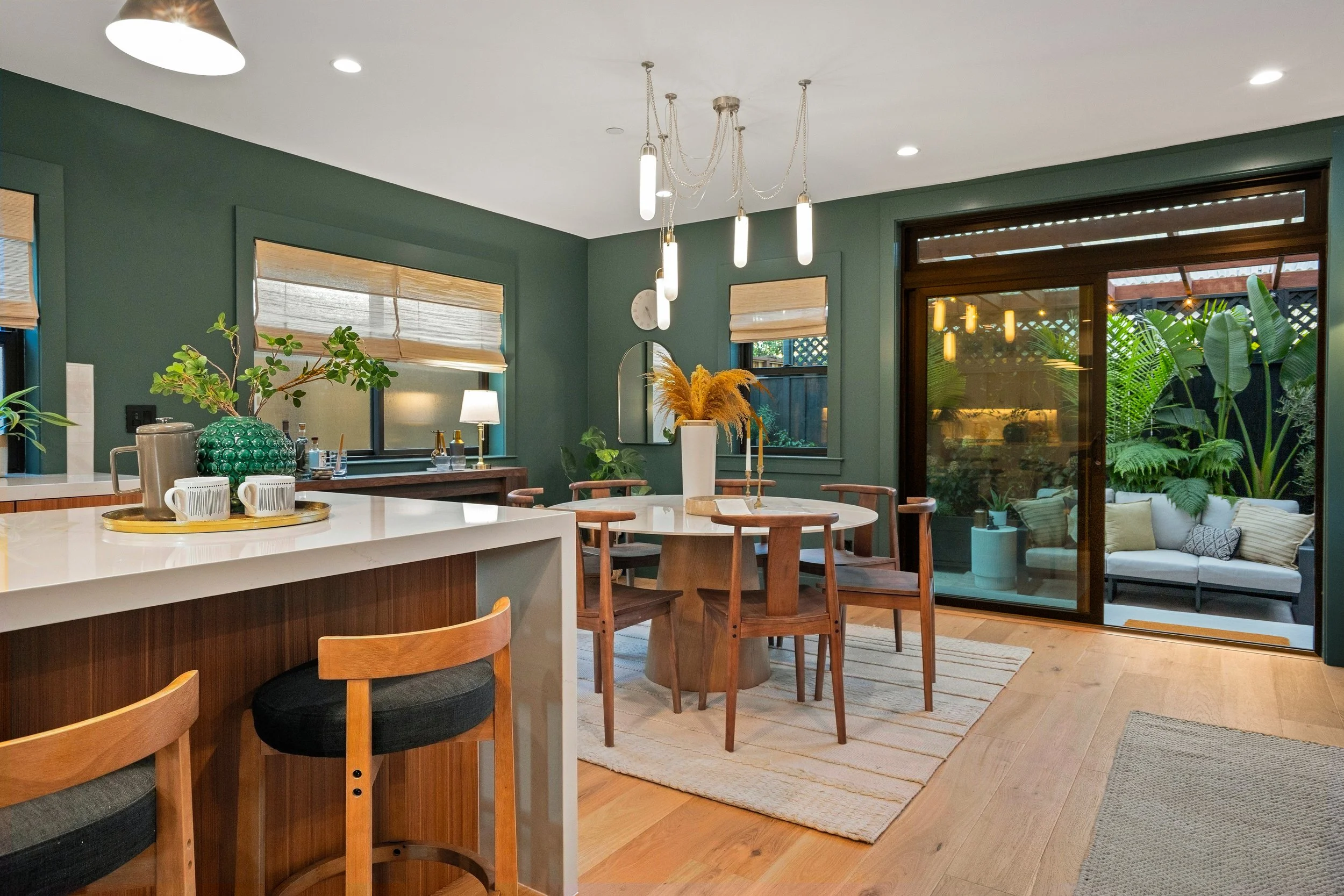 A modern kitchen and dining area with green walls, wooden furniture, pendant lights, and large windows and sliding glass door leading to an outdoor patio with garden furniture and lush greenery.