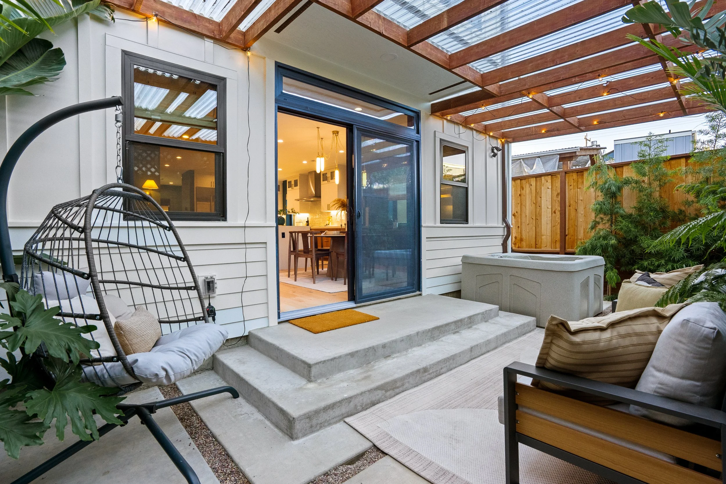 Back patio of a house with steps leading to a sliding glass door, outdoor furniture, plants, and a wooden pergola with string lights.