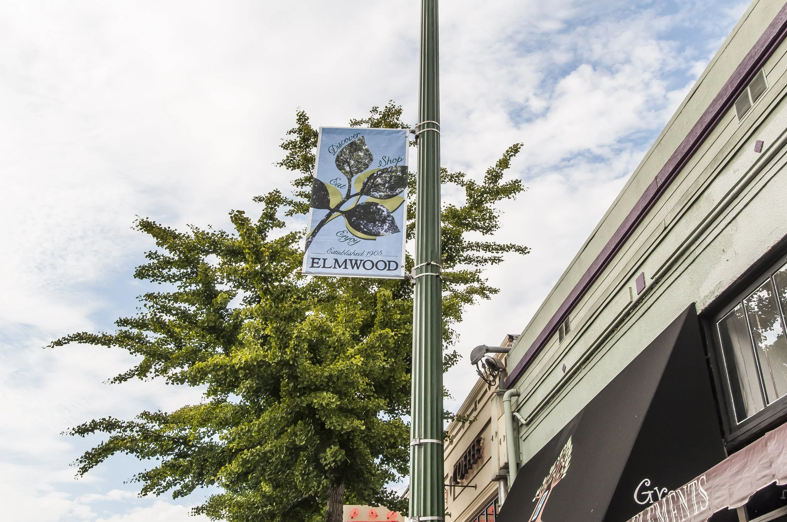 A street scene with a light green storefront, a tall lamppost with a decorative banner, green trees, and partly cloudy sky.