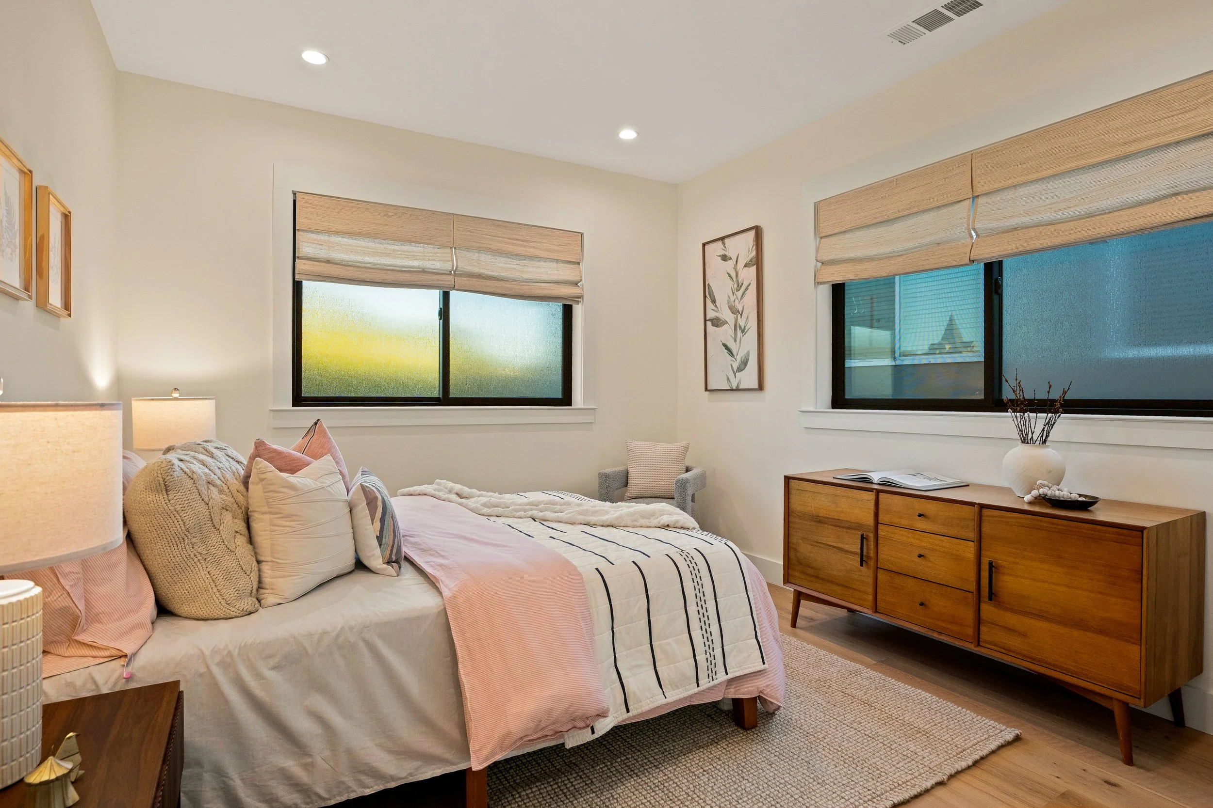 A bedroom with a bed, pillows, and blanket, two windows with beige shades, and a wooden dresser with a decorative vase on top.