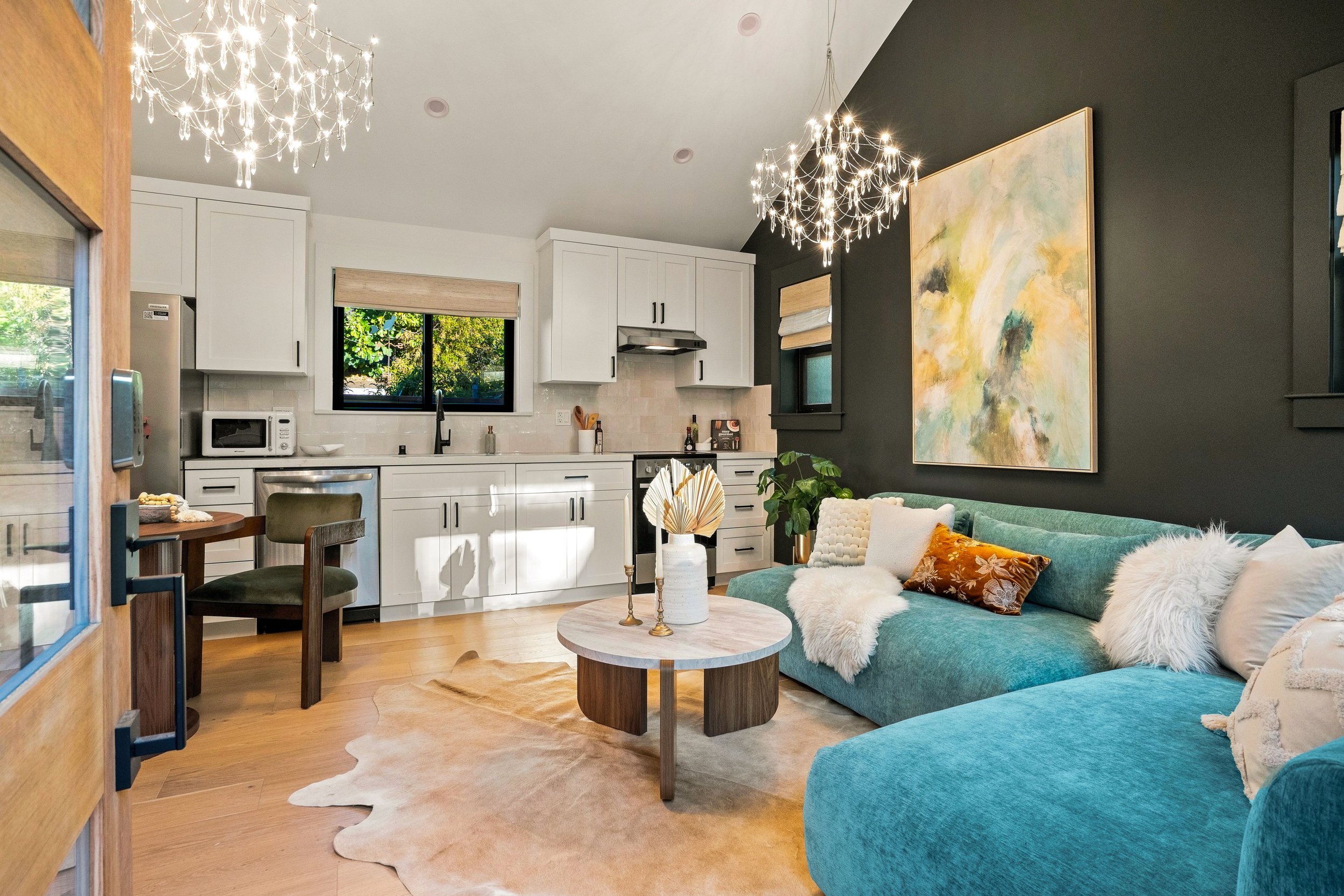 A modern open-concept living room and kitchen with white cabinets, a black wall, and contemporary decor. Decor includes plush teal couches with decorative pillows, a round marble coffee table, and wall art. Warm lighting is provided by two chandelier