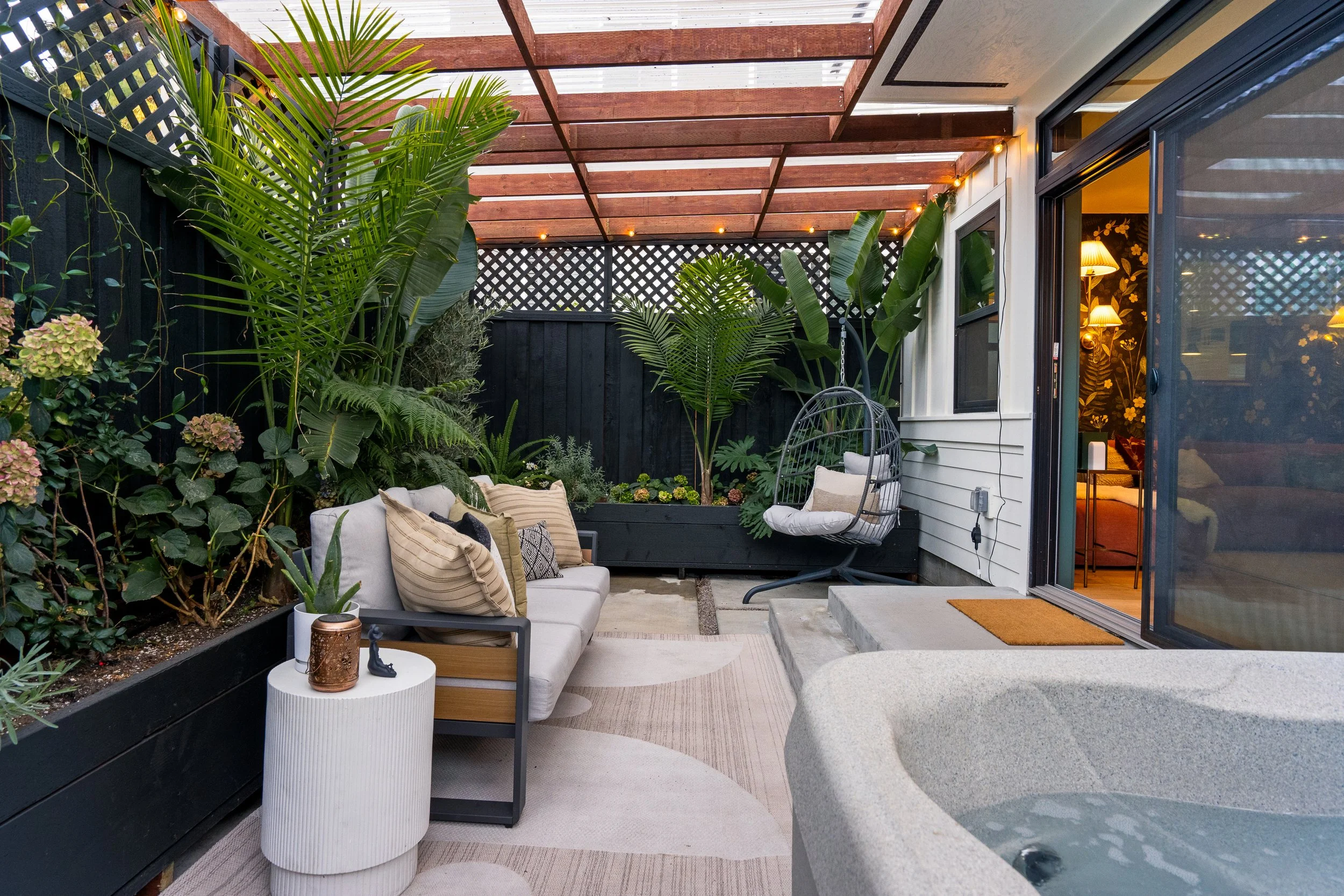 A cozy outdoor patio area with a white sofa with striped cushions, a small white side table with a candle holder, lush green tropical plants along the fence, a hanging egg chair, and a sliding glass door leading inside a warmly lit room. Wood slat pe
