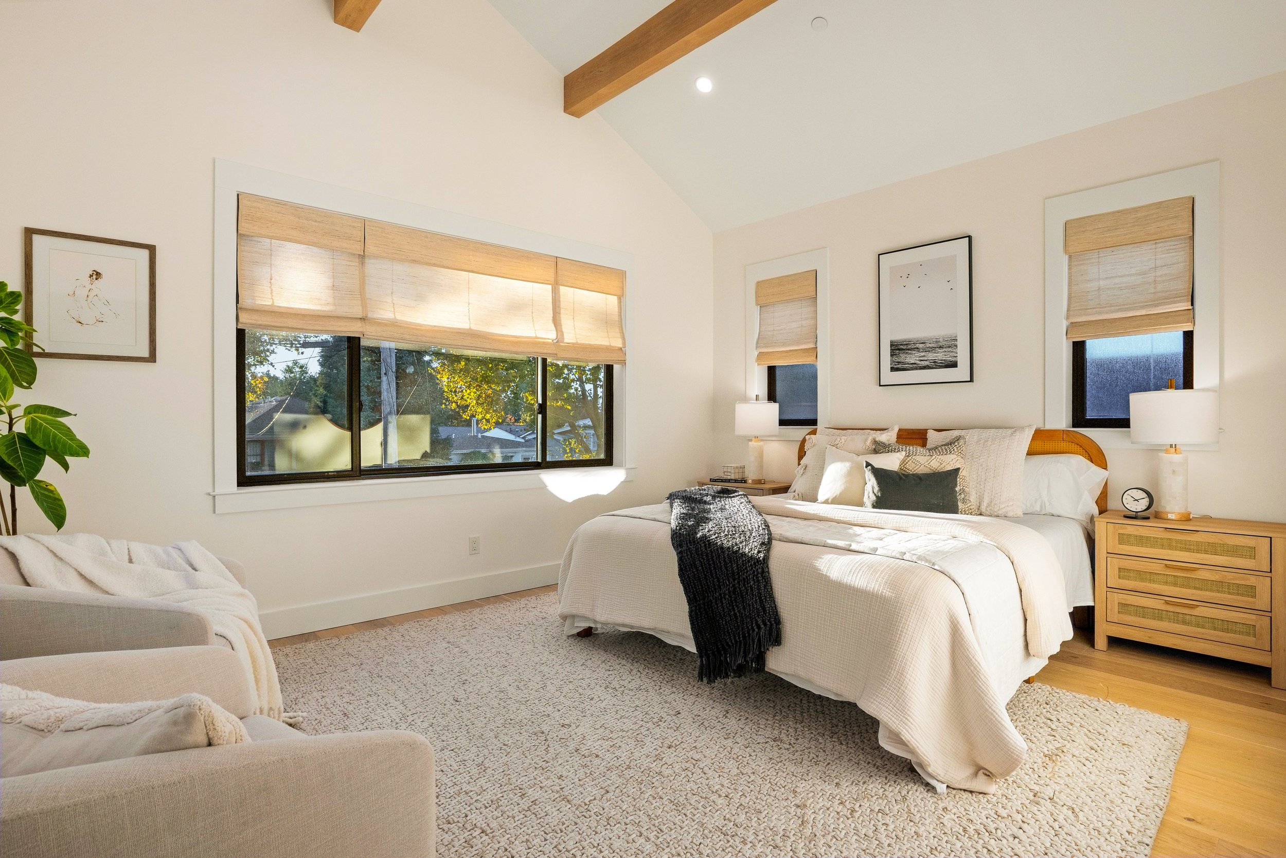 A cozy bedroom with a large bed, neutral-colored bedding, and wooden nightstands with white lamps. There are three windows with bamboo shades, framed artwork on the walls, and a beige carpet.