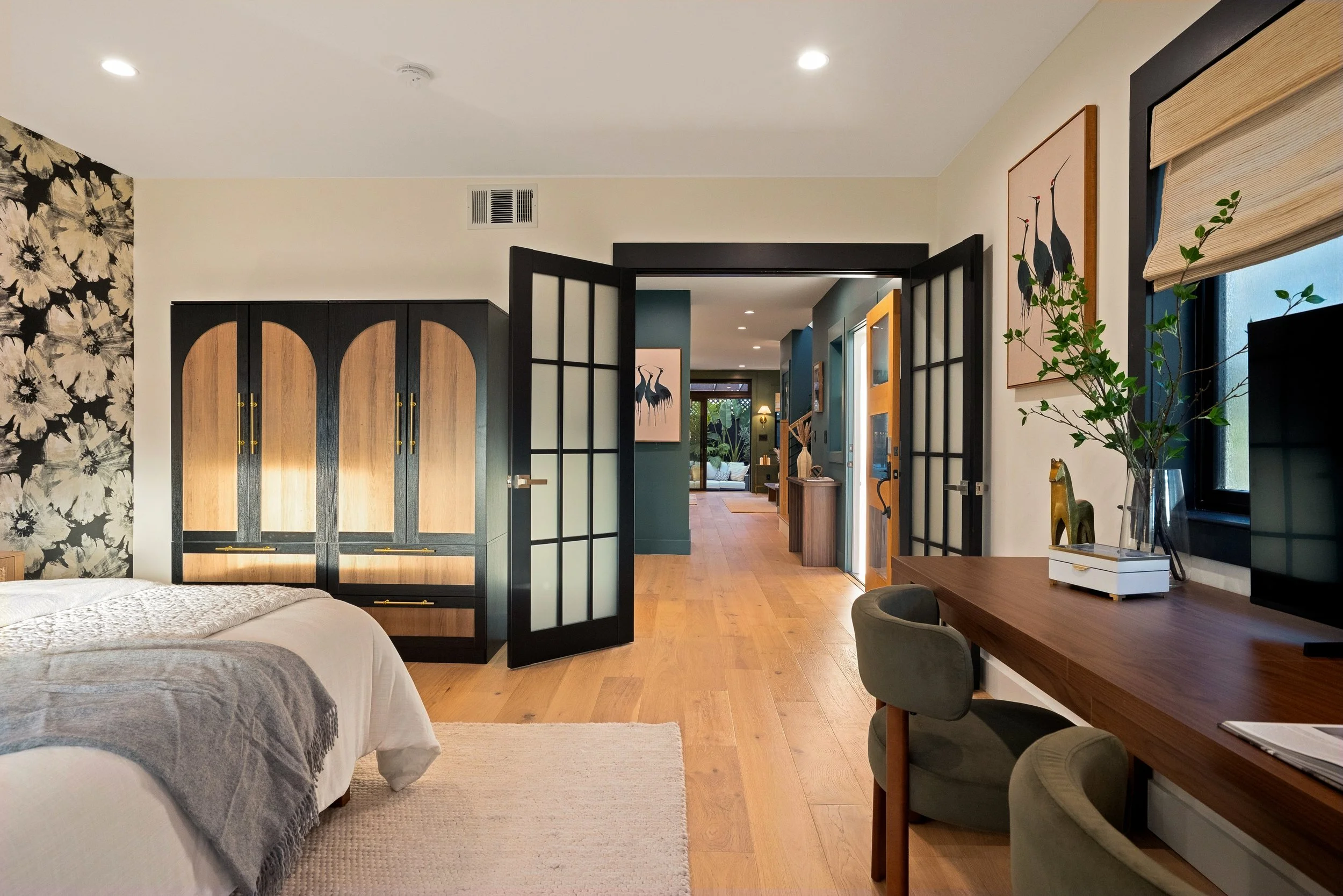 Interior view of a modern bedroom with wooden furniture, black and wood wardrobe, a glass room divider, plants, artwork of cranes, and a wooden desk with chairs.