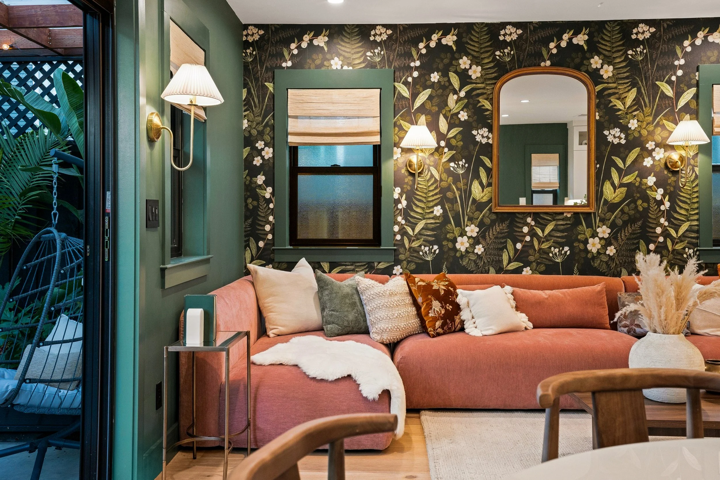 Living room with a pink sectional sofa decorated with various pillows, a white furry throw, and a beige vase with pampas grass. The wall behind is covered with dark floral wallpaper, a large mirror with a wooden frame, and two wall-mounted lamps with