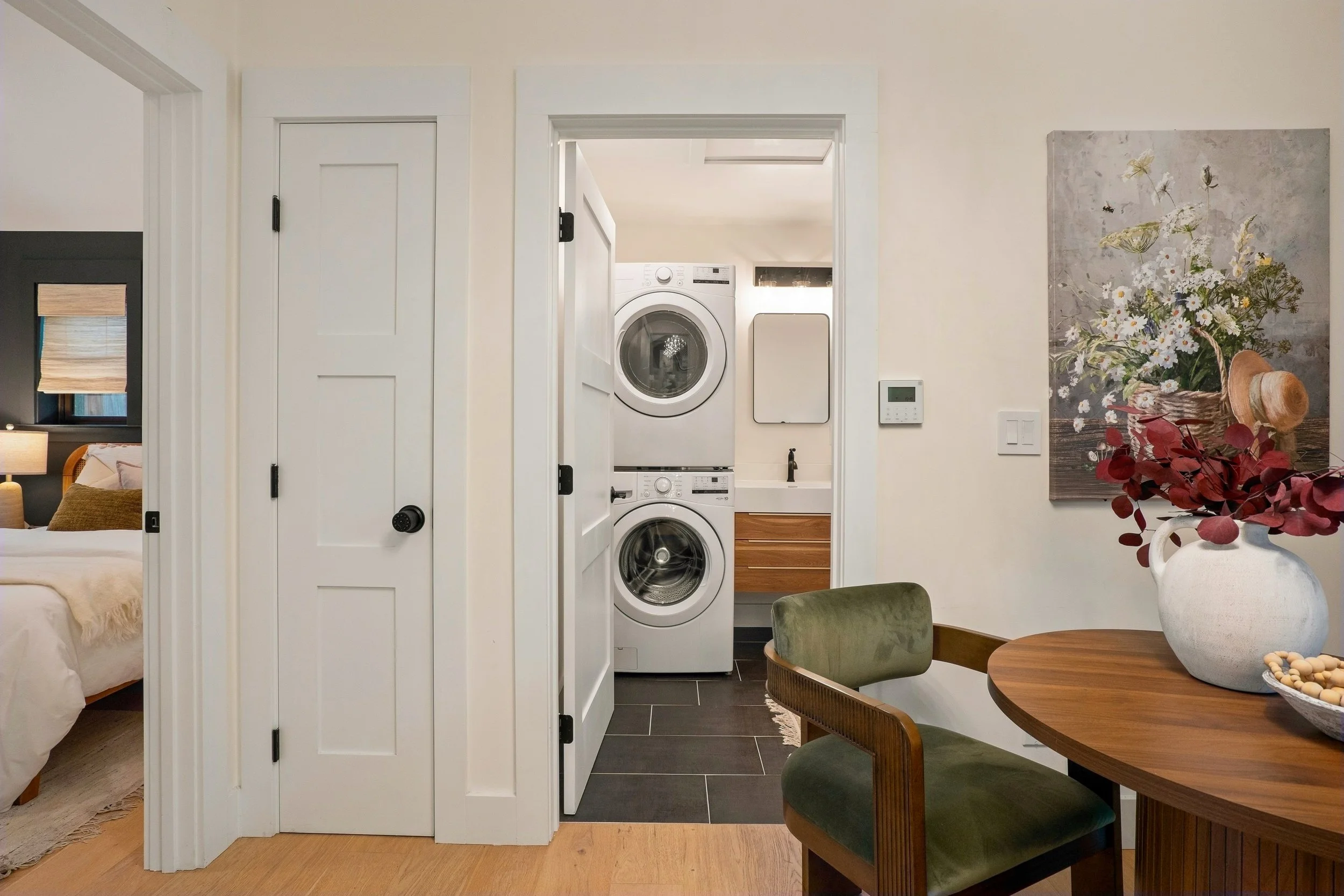 View of laundry area with stacked washer and dryer behind a white door, adjacent to a dining table with green upholstered chairs and a white vase with flowers on the table.