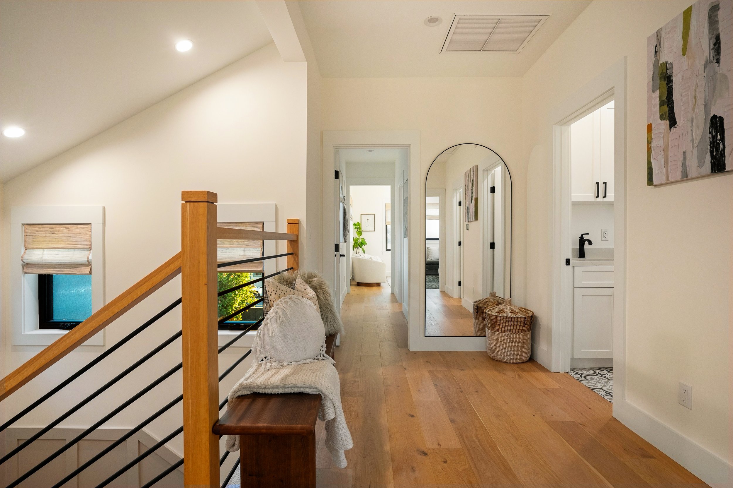 Interior hallway of a modern home with white walls, hardwood floors, a mirror, abstract artwork, wicker baskets, a window, and a staircase with a wooden handrail.