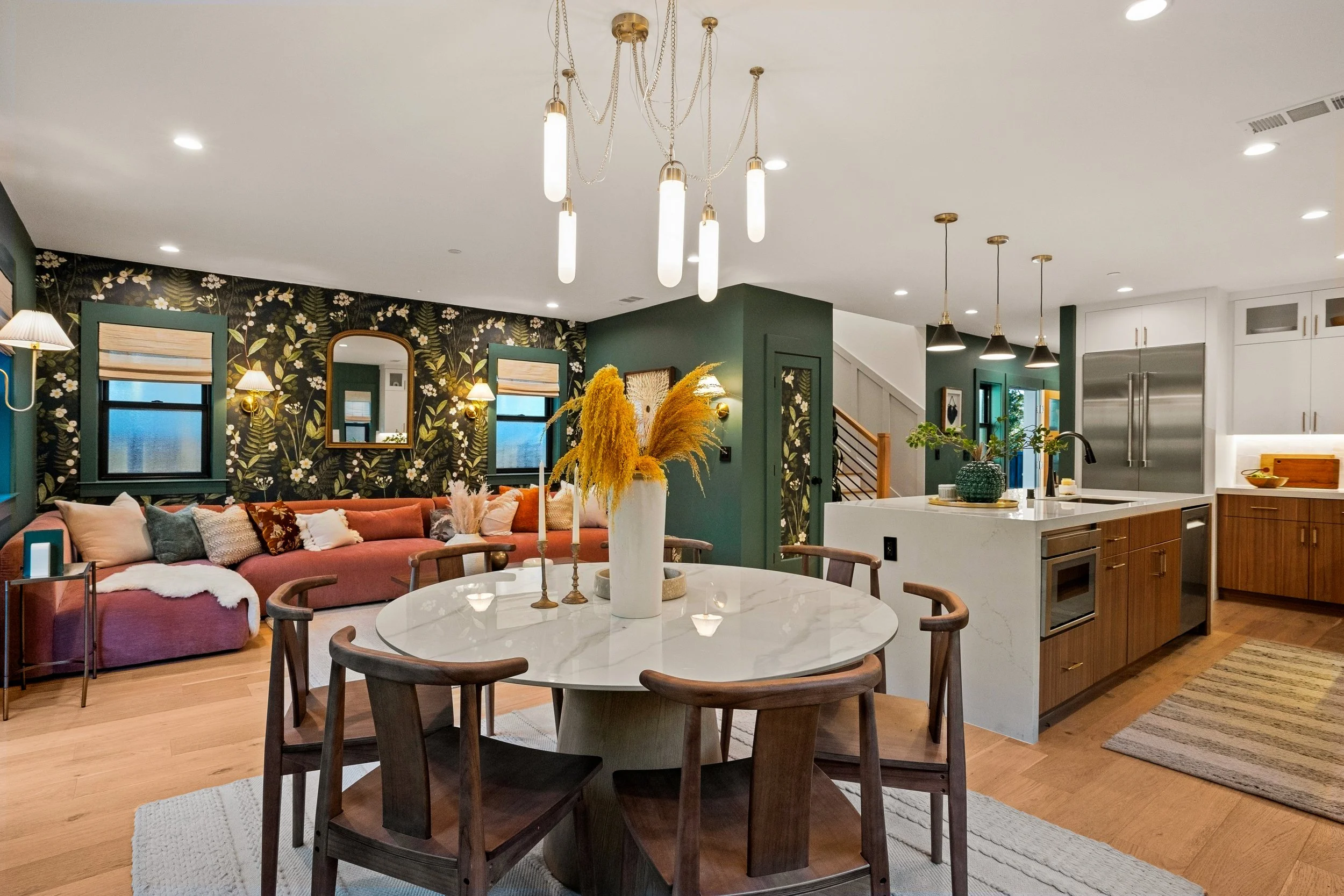 A living room and kitchen area with a green accent wall, dark floral wallpaper, a pink sectional sofa, and a round dining table with wooden chairs. Modern lighting fixtures hang from the ceiling, and there are decorative plants and mirrors.