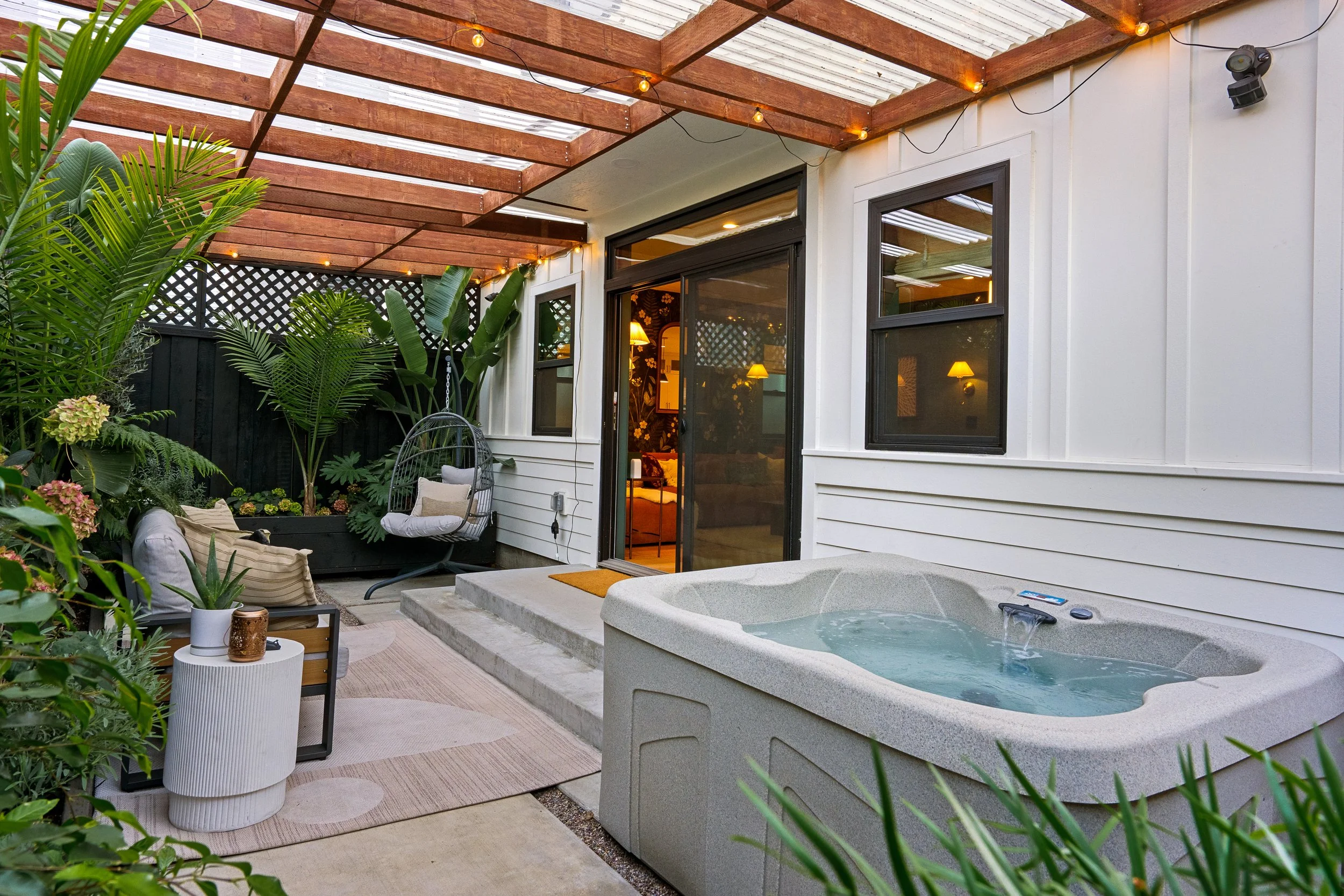 A cozy backyard patio area featuring a hot tub, cushioned chairs, lush green plants, and string lights under a wooden pergola, viewed through a sliding glass door.