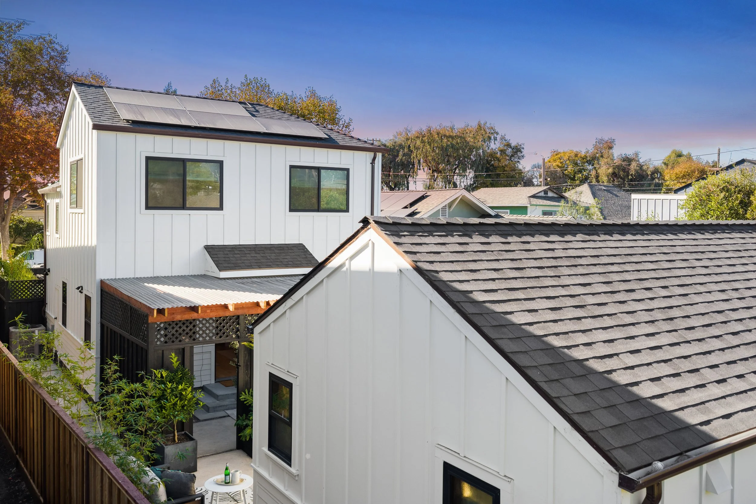 View of a modern white house with solar panels on the roof, surrounded by trees with autumn foliage, neighboring houses, and a backyard with lounge furniture and a small table.