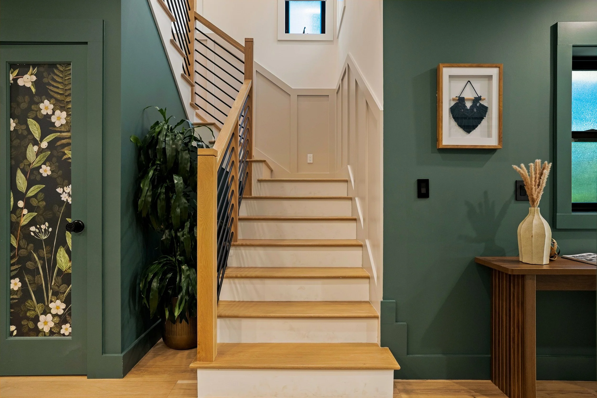 Interior view of a staircase with wooden steps and black metal railing, against a green wall with framed floral and black macrame art, a tall potted plant, and a wooden table with a vase containing dried grasses.